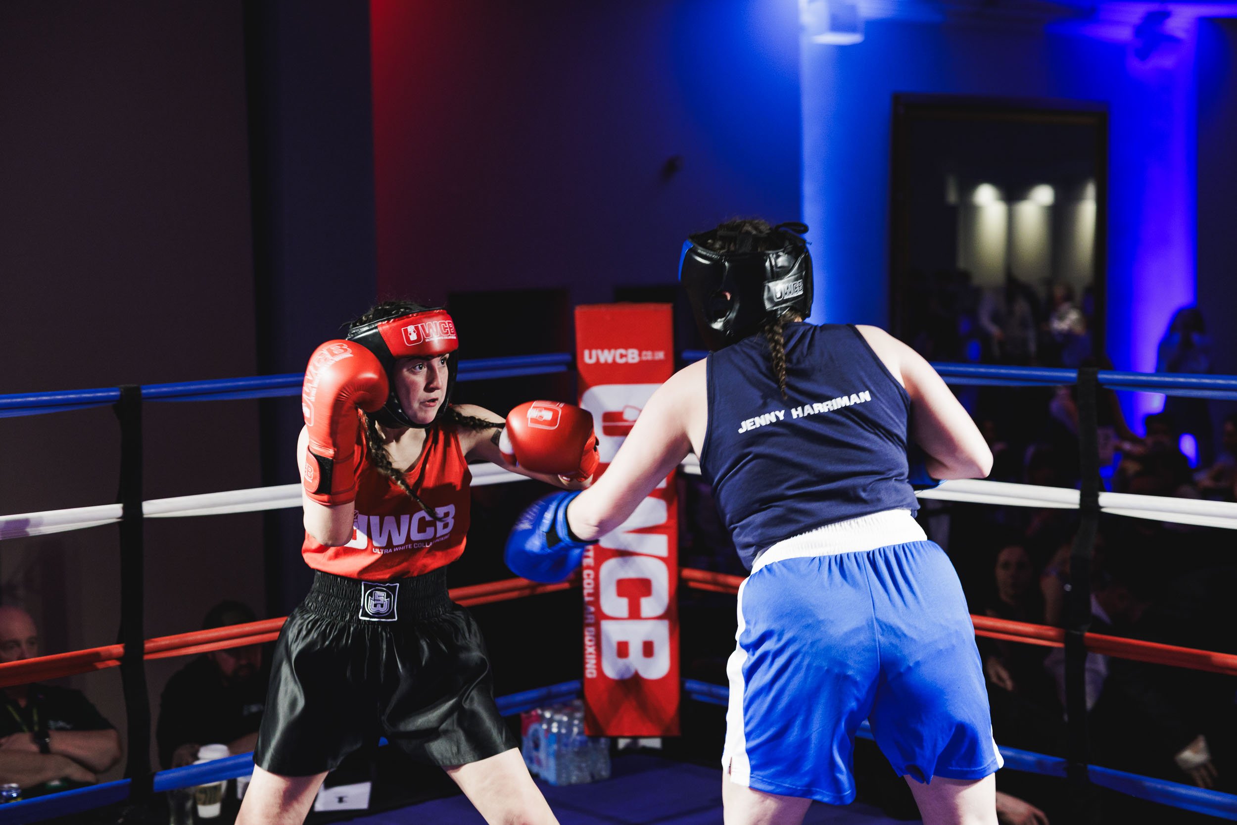 Two women boxers sparring in a boxing ring during a match, wearing protective headgear, gloves, and athletic clothing. One is in red gloves and top, the other in blue, with the name "Jenny Harriman" visible on her shirt.