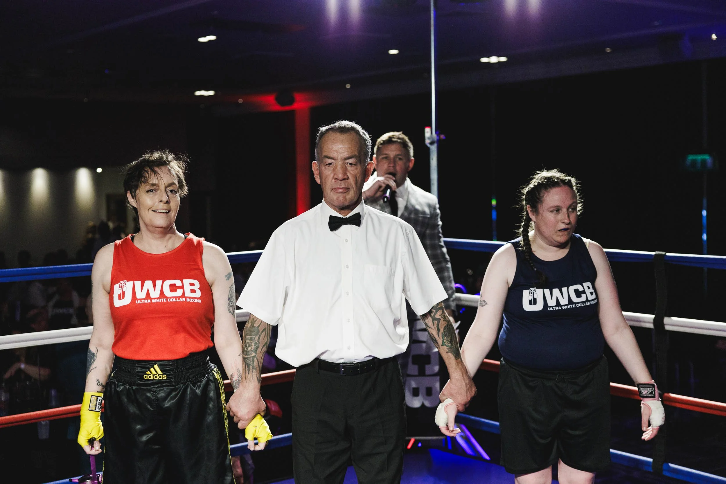 Two female boxers, one in a red tank top and the other in a blue tank top, hold hands in a boxing ring with a referee between them. A man with a microphone stands behind the referee in a casino or event hall setting.