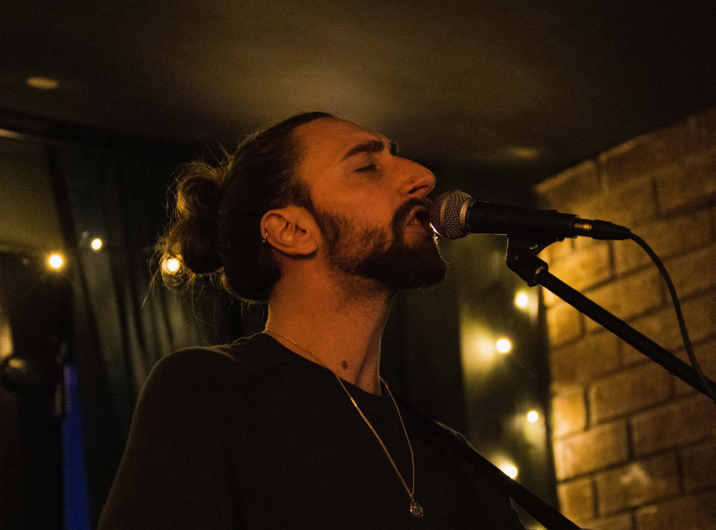A man with a beard and long hair tied in a bun singing into a microphone at an indoor venue with warm lighting and a brick wall in the background.