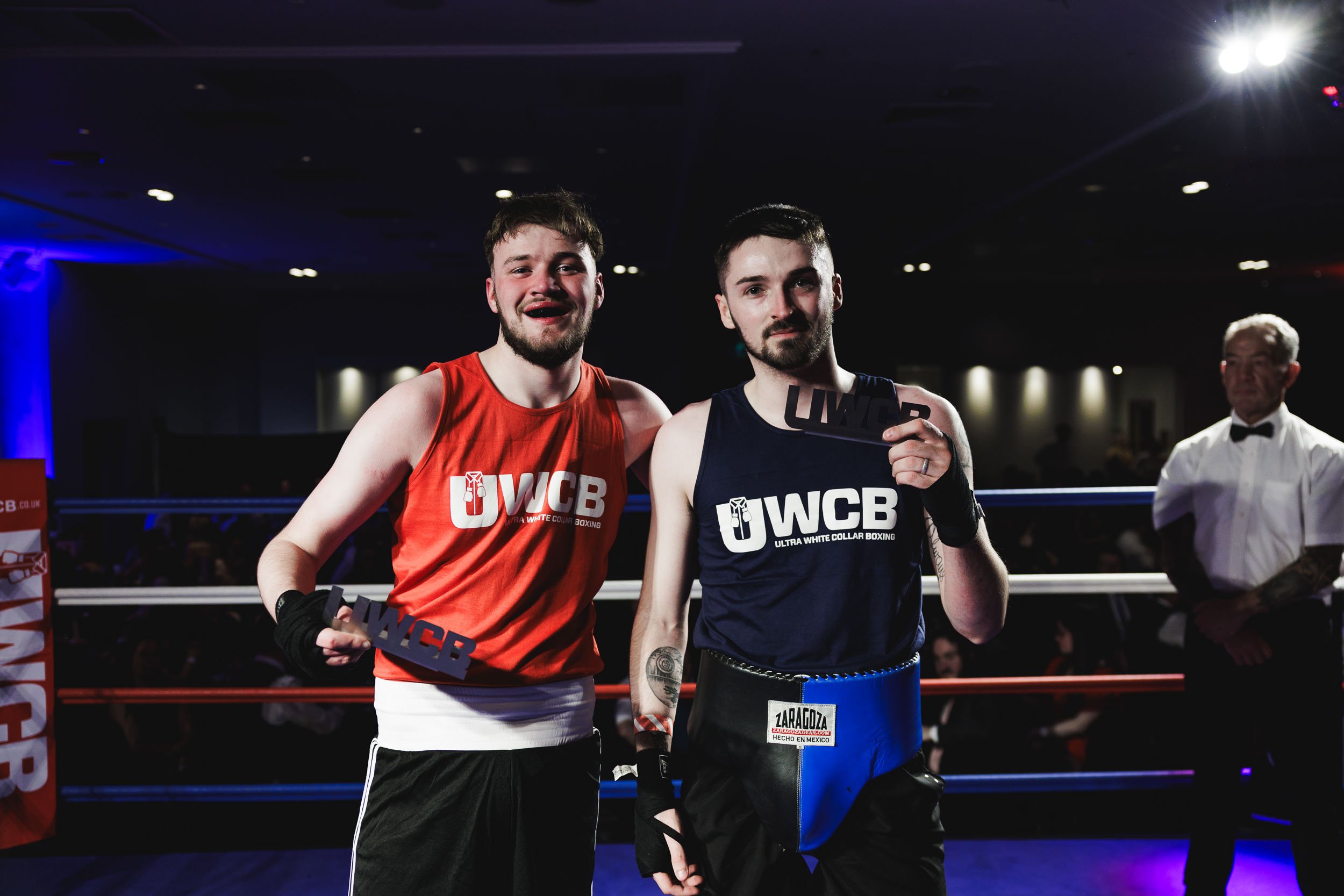Two male boxers in a boxing ring holding trophies, wearing red and blue trunks with 'U WCB' logos, with a referee and audience in the background.