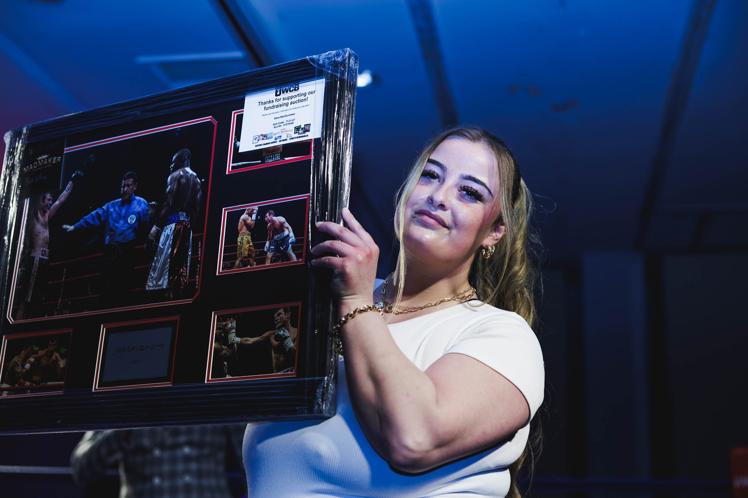 A woman with wavy blonde hair holding a framed collage of photographs from a boxing event, smiling slightly, dressed in a white top with gold jewelry, in a dimly lit environment.