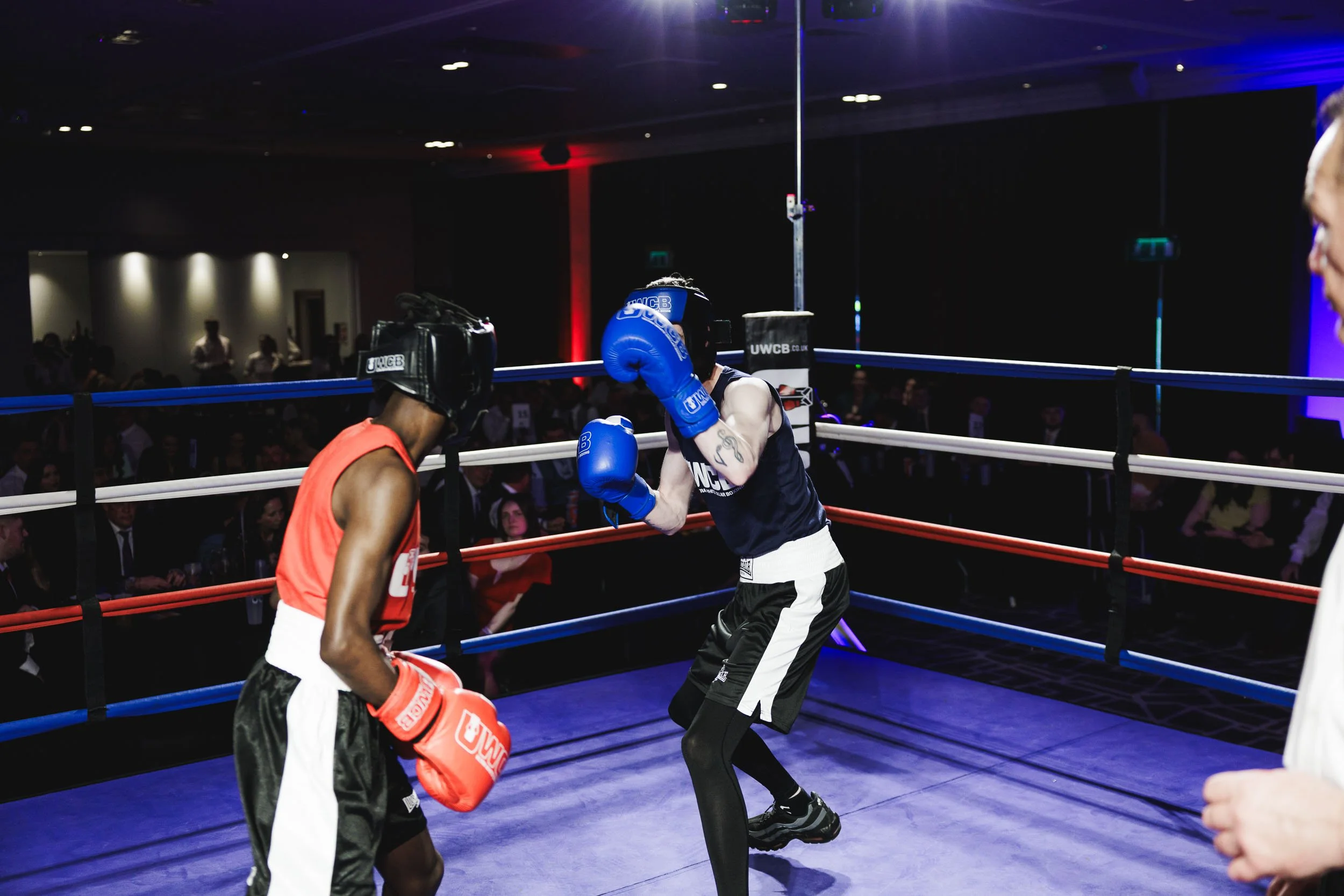 Two young boxers wearing protective gear sparring in a boxing ring, with an audience watching in the background.