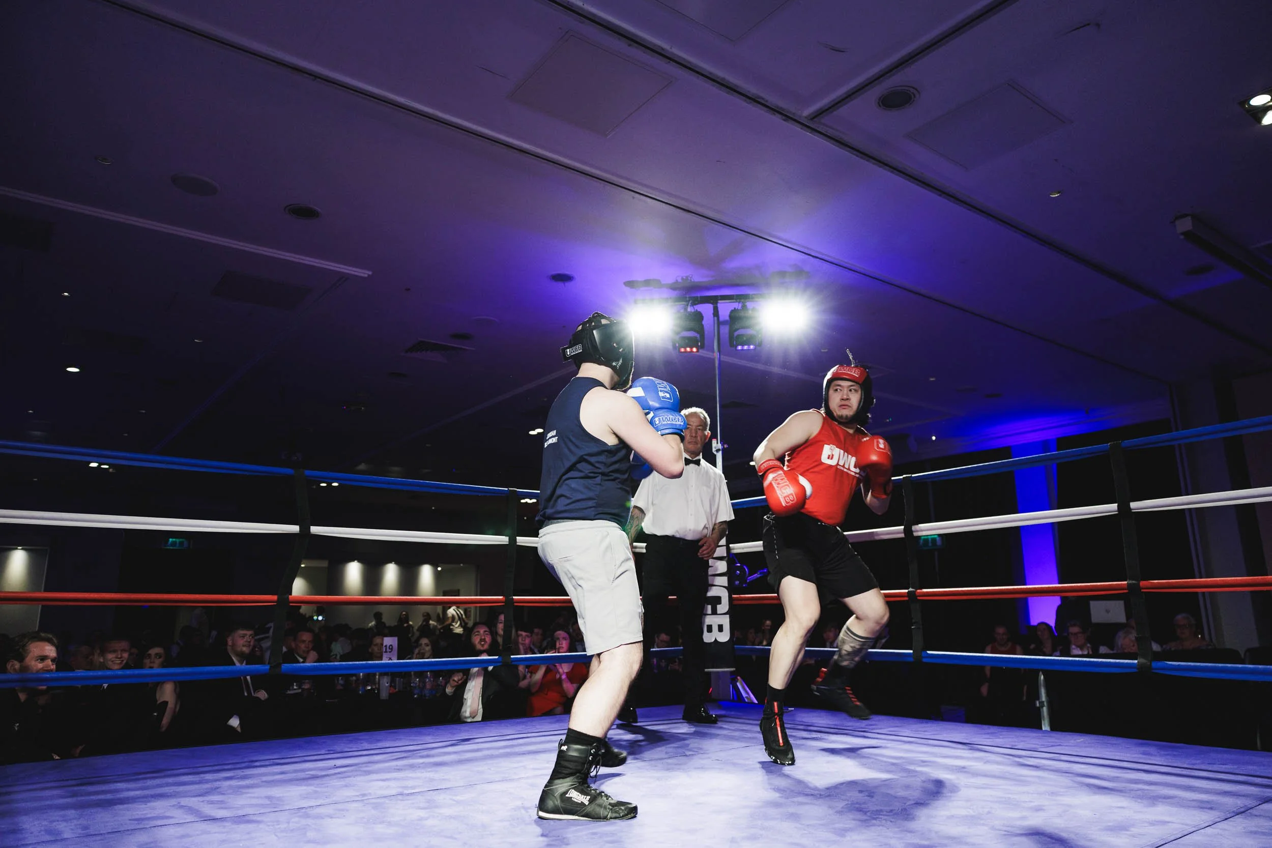 Two men competing in a boxing match inside a boxing ring with an audience watching, one wearing red gear and the other blue, with a referee observing.
