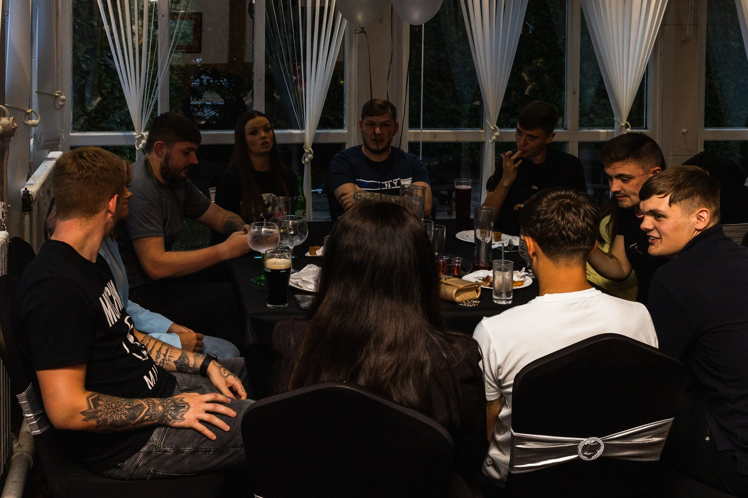 Group of young people sitting around a dining table in a dimly lit indoor setting during evening.