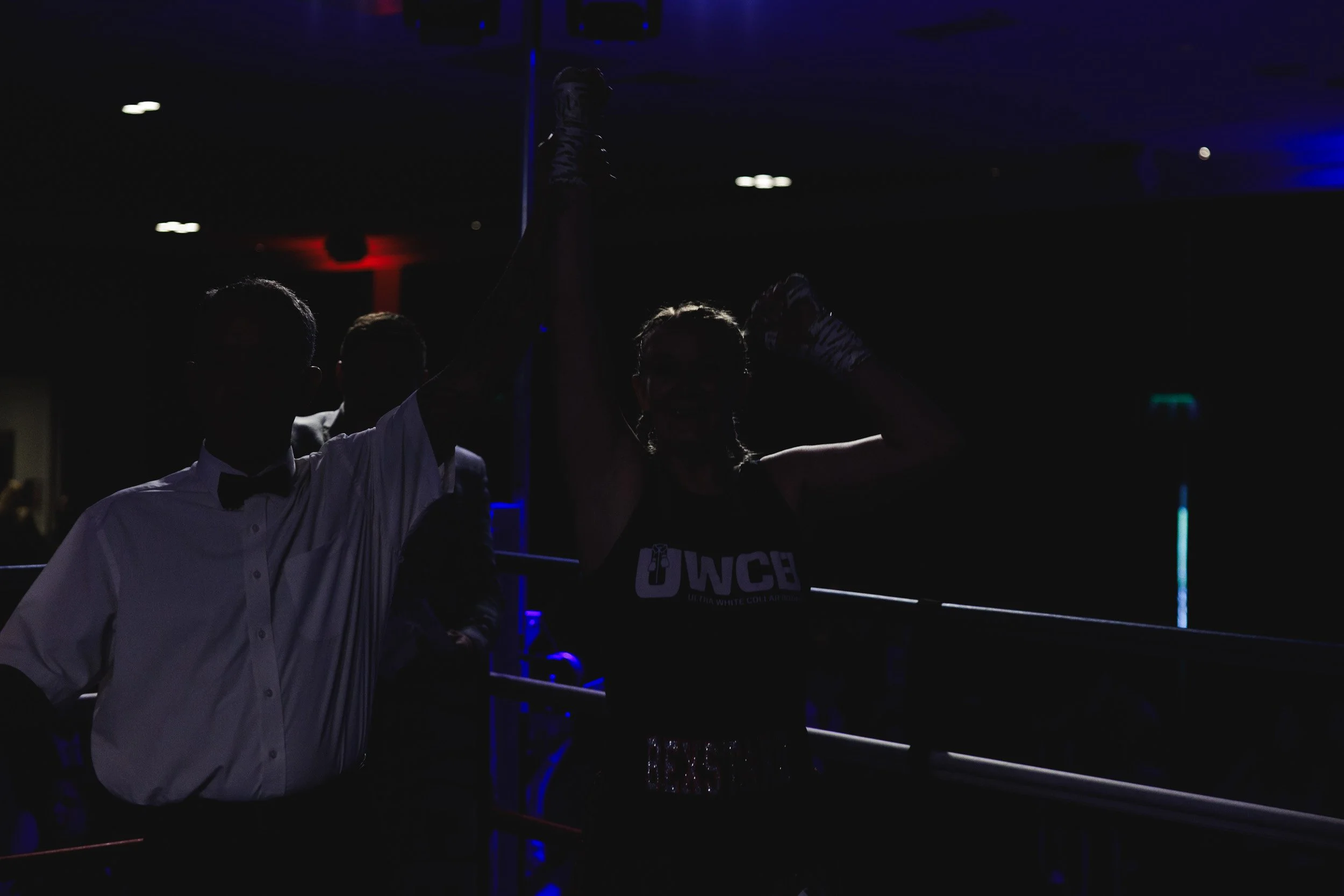 A boxing match winner, a woman with a sports bra and gloves, raising her arm in victory while standing in a boxing ring with a man in a white shirt and bow tie, in a dark-lit environment with some blue and red lighting.
