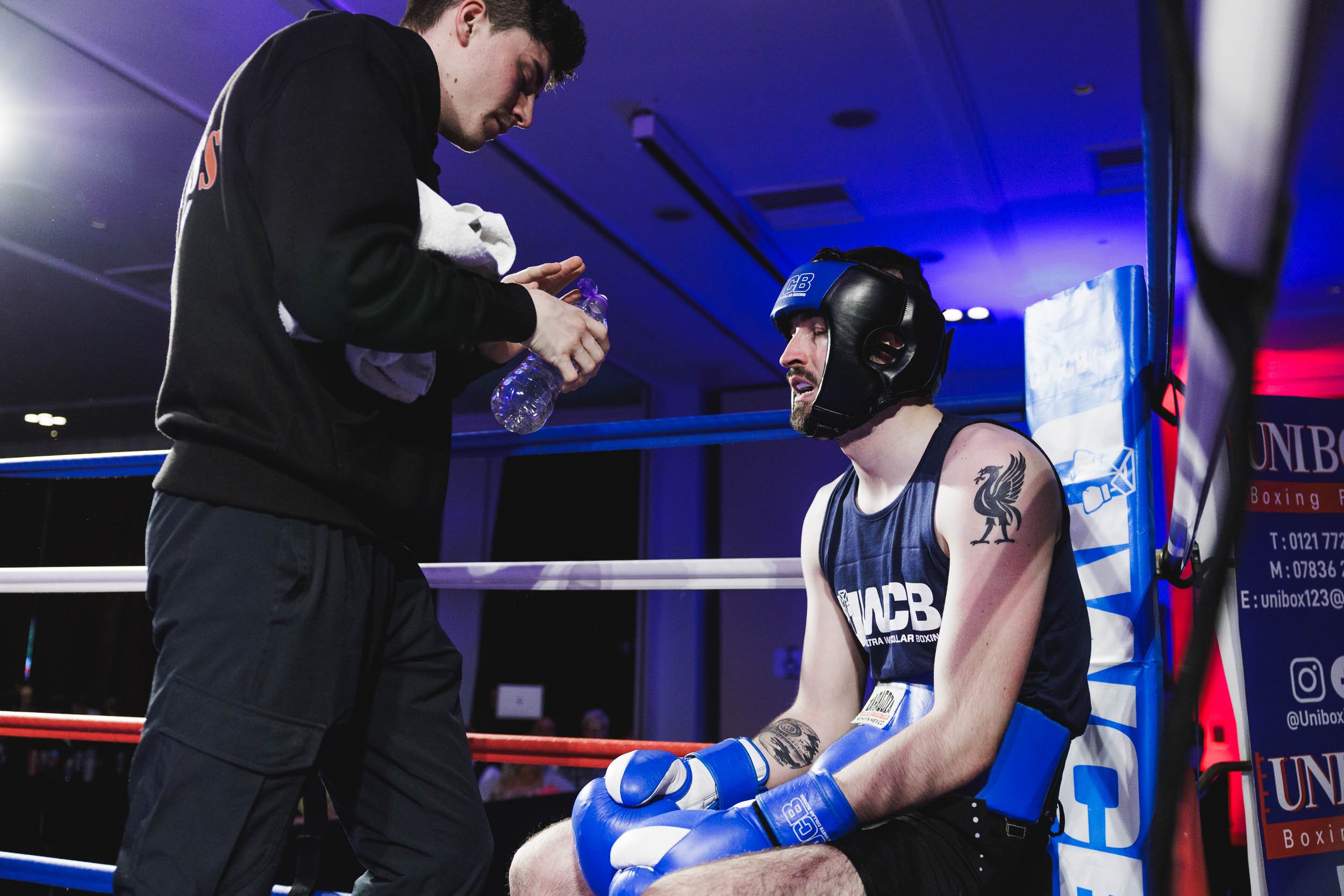 A boxer sitting on the corner of a boxing ring, wearing a black helmet and blue gloves, looks tired and injured. A team member is standing nearby, holding a water bottle and towel, preparing to help the boxer during a break in the match.