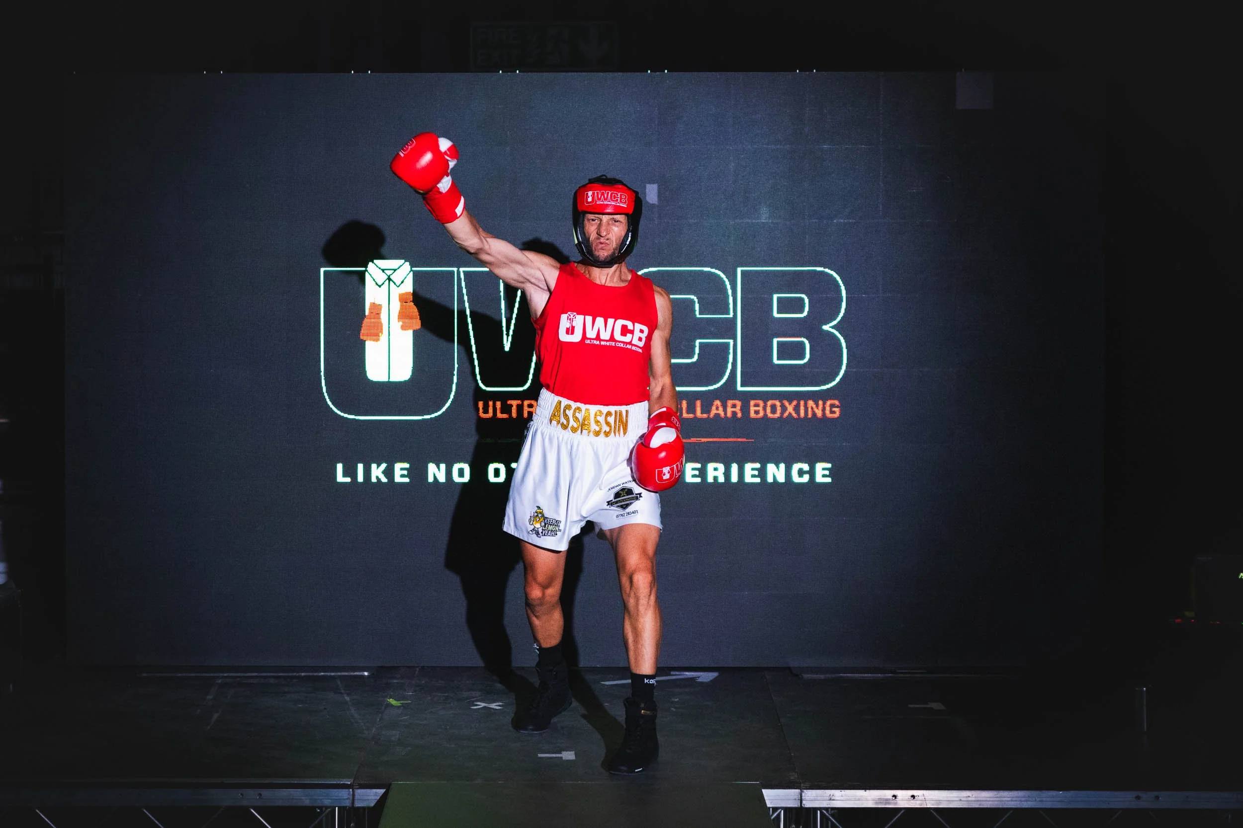 A male boxer in red training gear with red gloves and white shorts standing on stage with his right arm raised. Behind him is a large screen displaying a logo for ultra white collar boxing, with the text 'LIKE NO OTHER EXPERIENCE'.