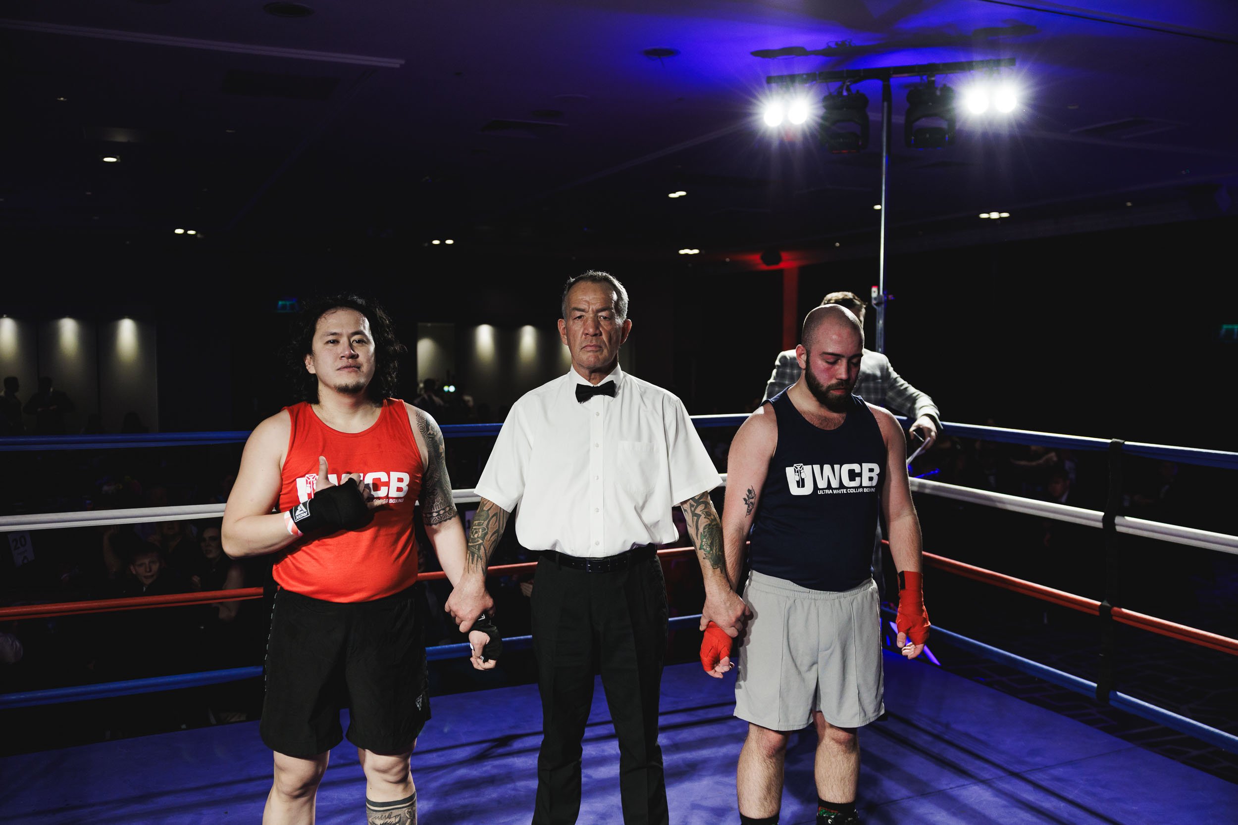 Two male boxers and a referee standing in a boxing ring, with the boxers holding hands, in an indoor arena with viewers.