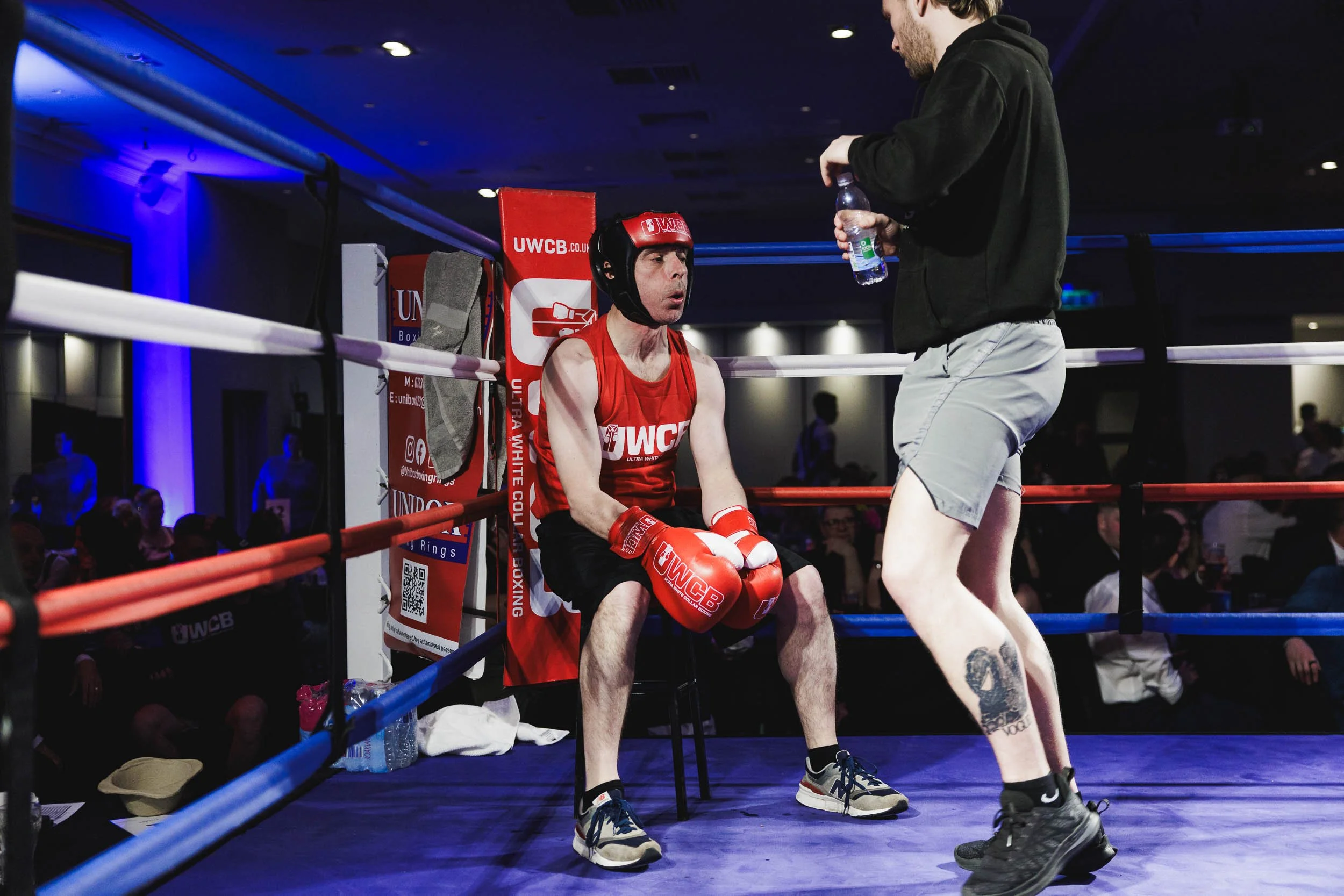 Boxer sitting on a stool inside a boxing ring, wearing red gloves, head protector, and tank top, talking to a man standing in front of him holding a water bottle.