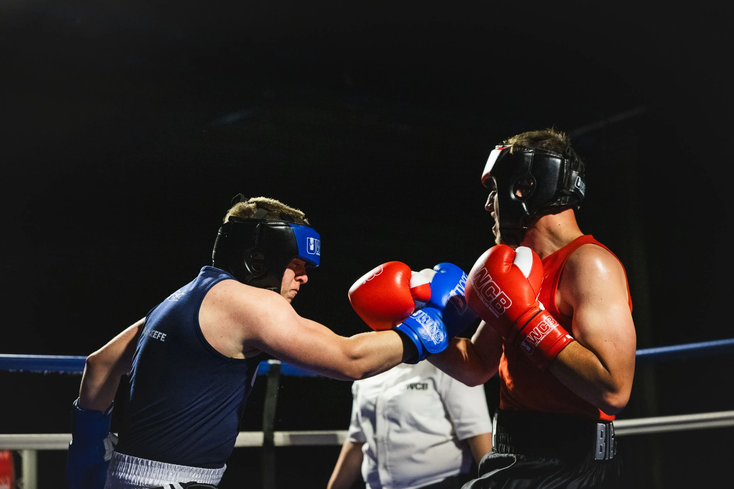 Two male boxers in boxing gloves and protective headgear sparring in a boxing ring, with a referee in the background.