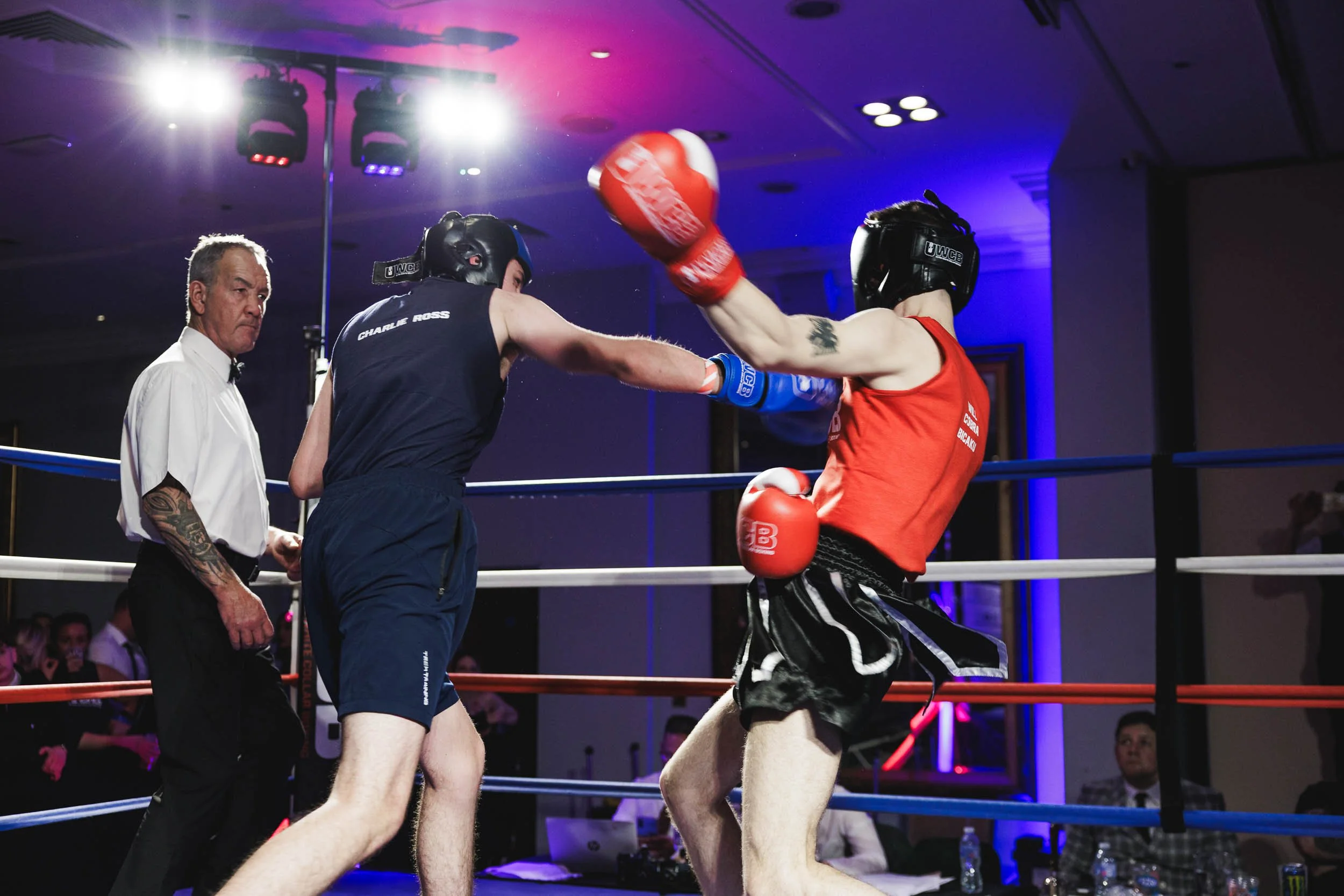Two young men wearing boxing uniforms and helmets fight in a boxing ring while a referee watches. One is throwing a punch, and the other is blocking.