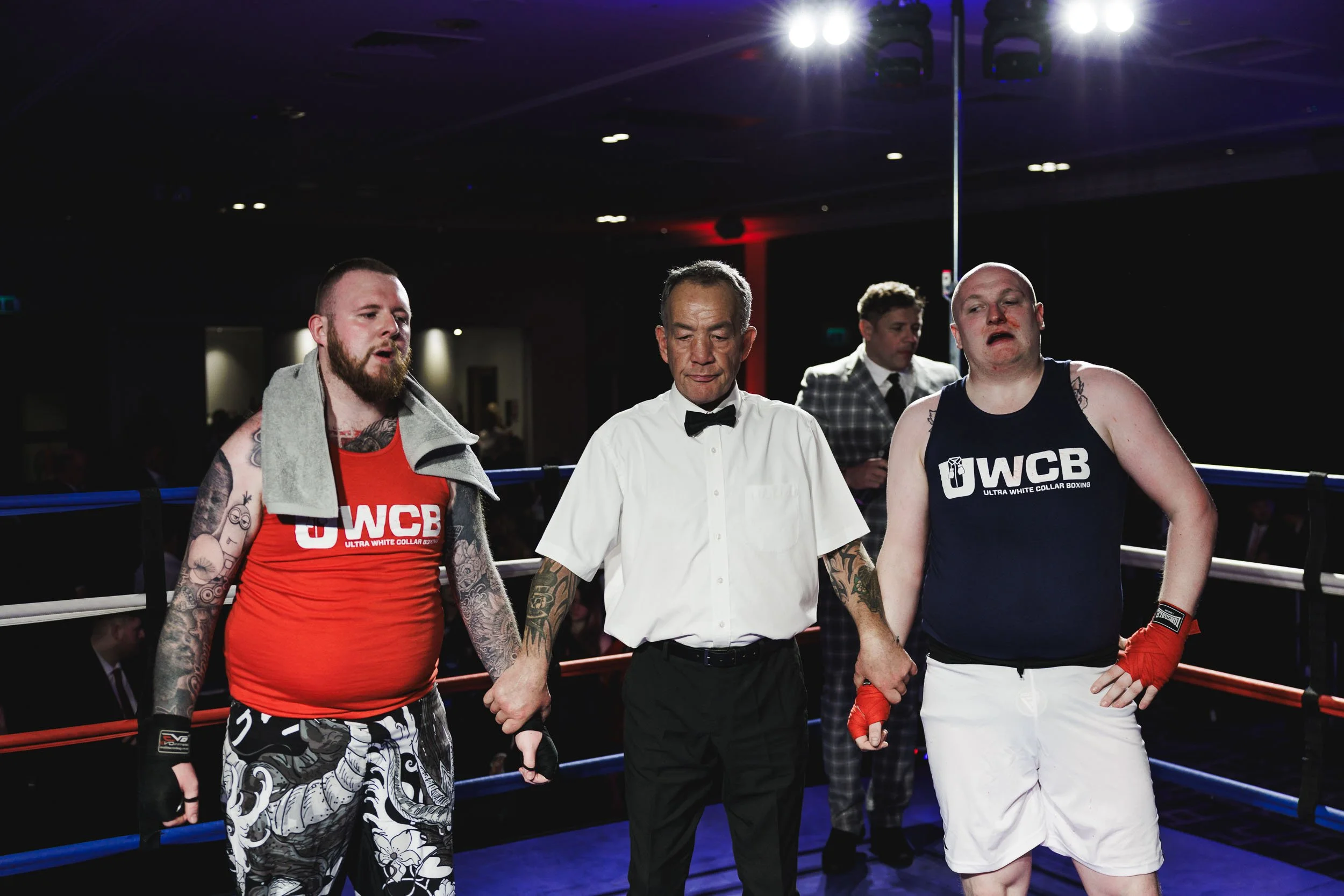 Two male boxers in boxing rings after a match, with a referee holding their hands together. The boxer on the left has tattoos, a red sleeveless shirt, and patterned shorts. The boxer on the right is wearing a black sleeveless shirt, white shorts, and