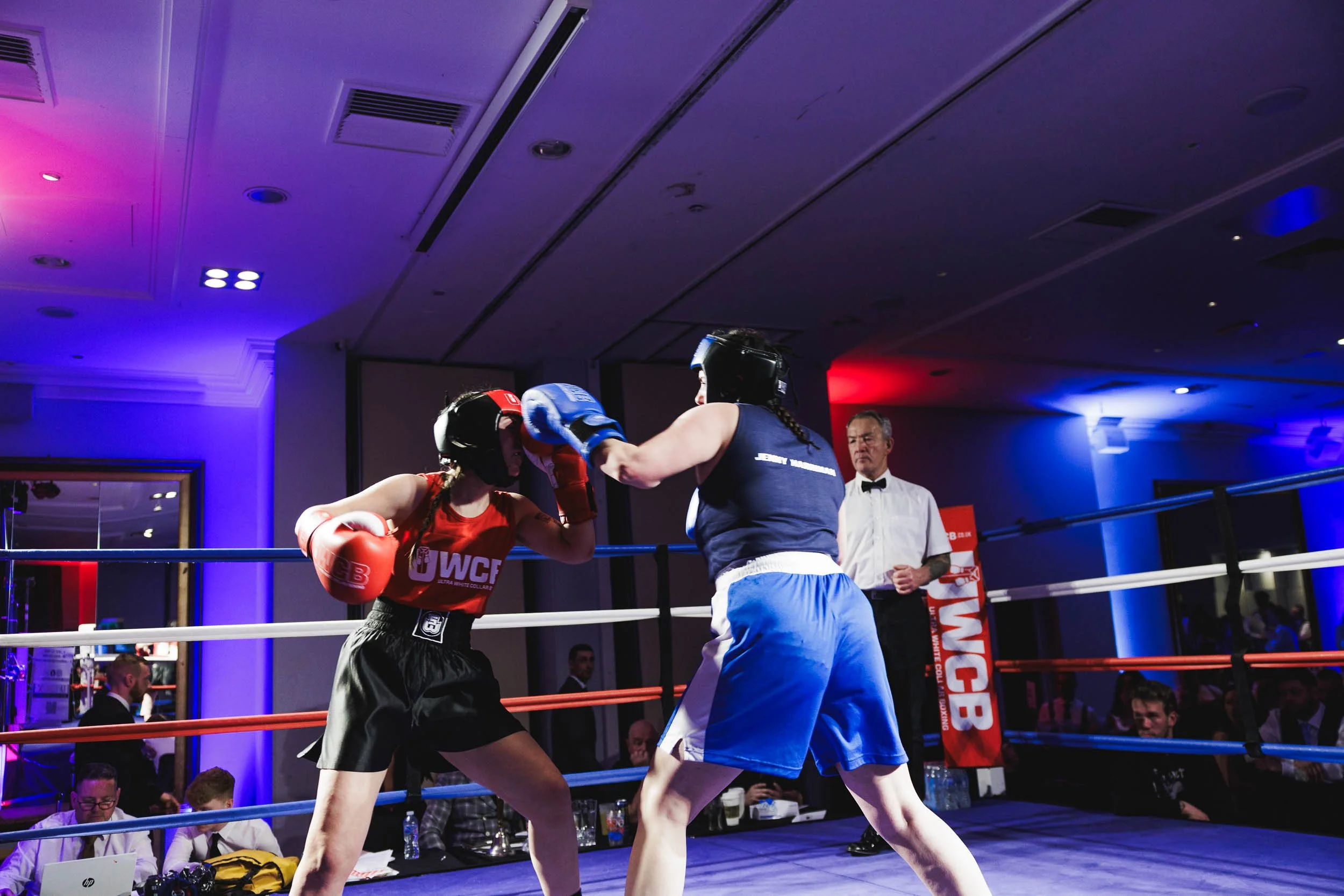 Two women boxers in a ring, one wearing red gloves and a red top, the other wearing blue gloves and blue shorts, sparring with a referee observing in the background.