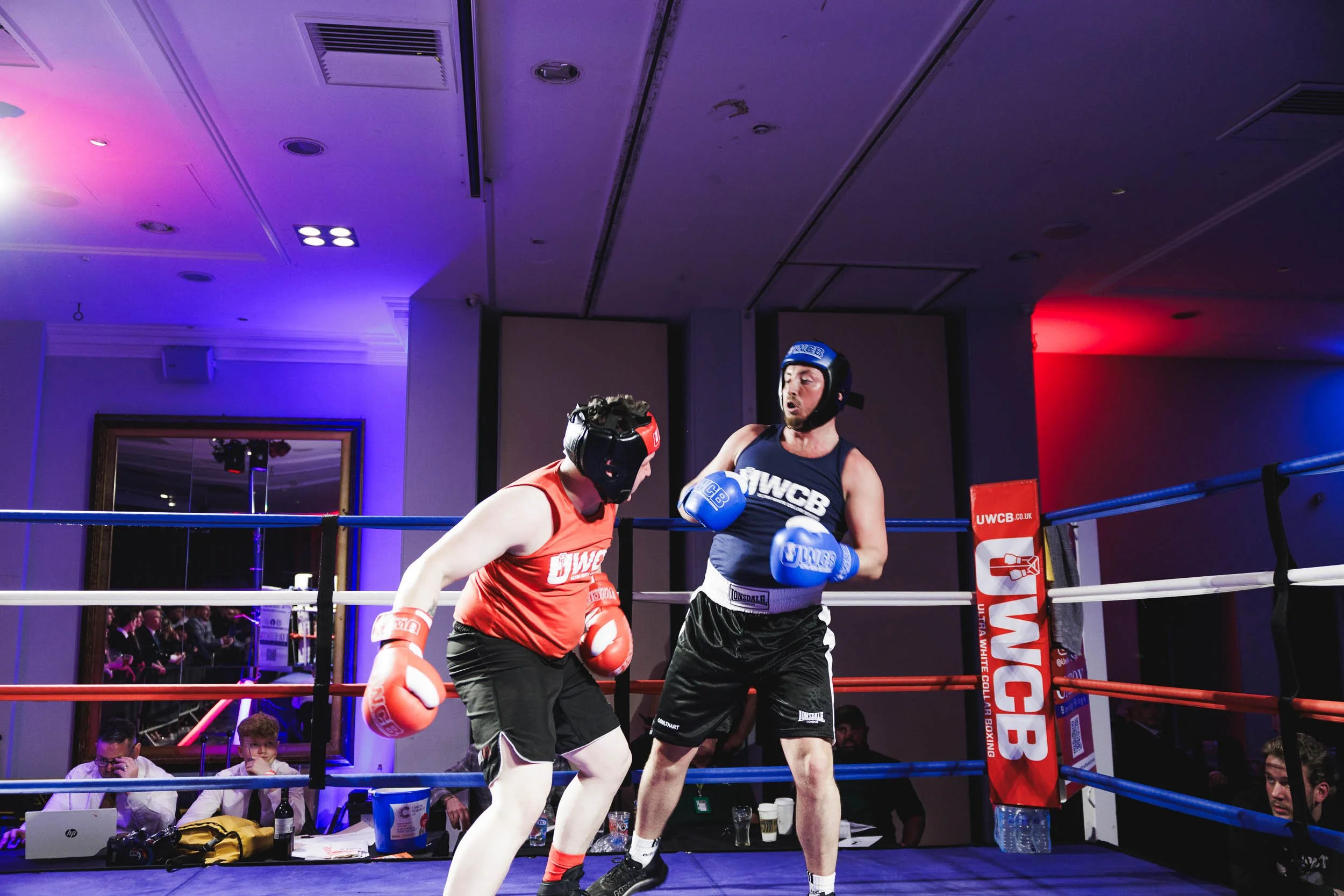Two amateur boxers in a ring, wearing protective headgear and gloves, engaged in a match with a referee observing closely.