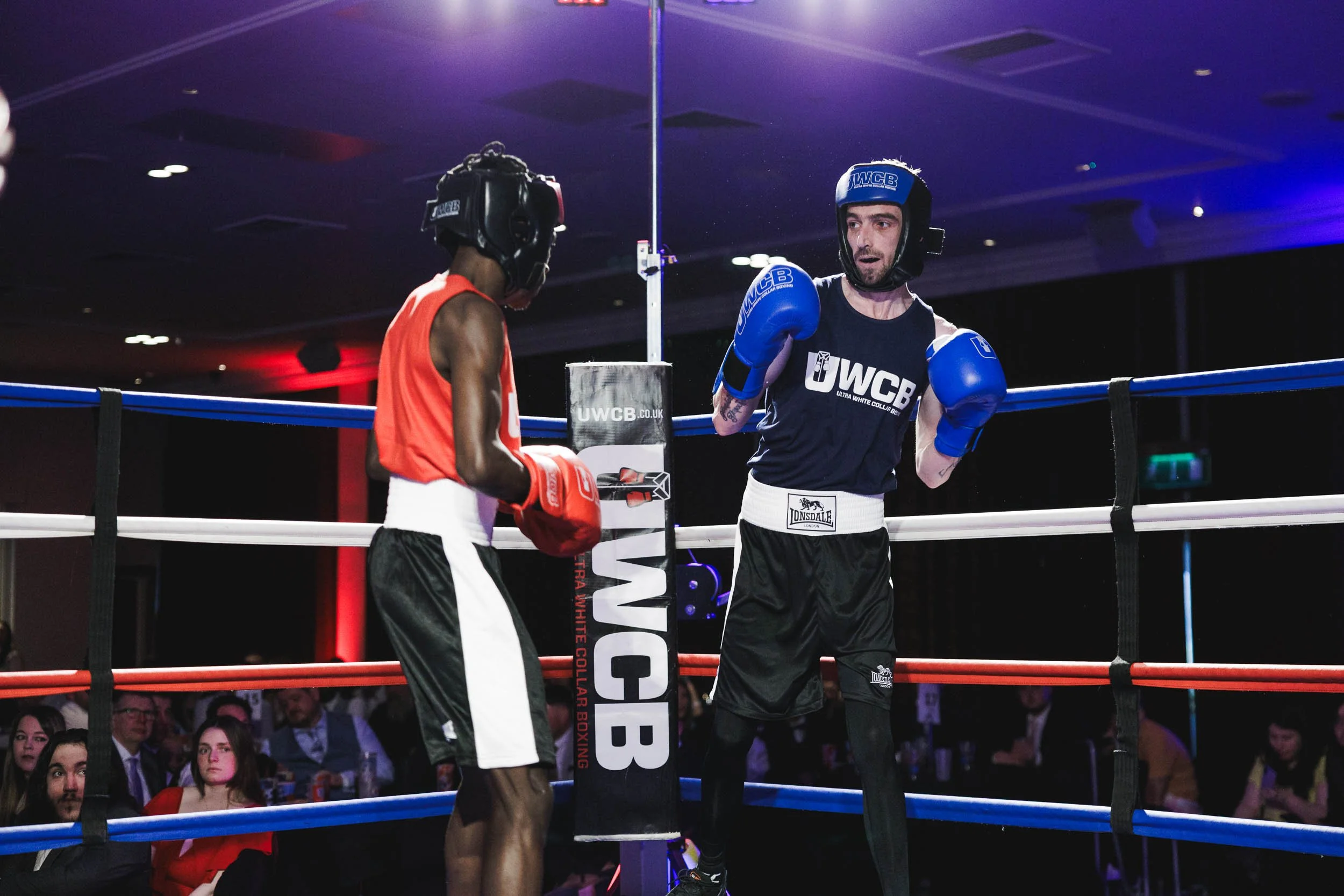 Two amateur boxers in boxing ring, one in red gear and one in blue gear, exchanging punches during a match, with audience seated around the ring.