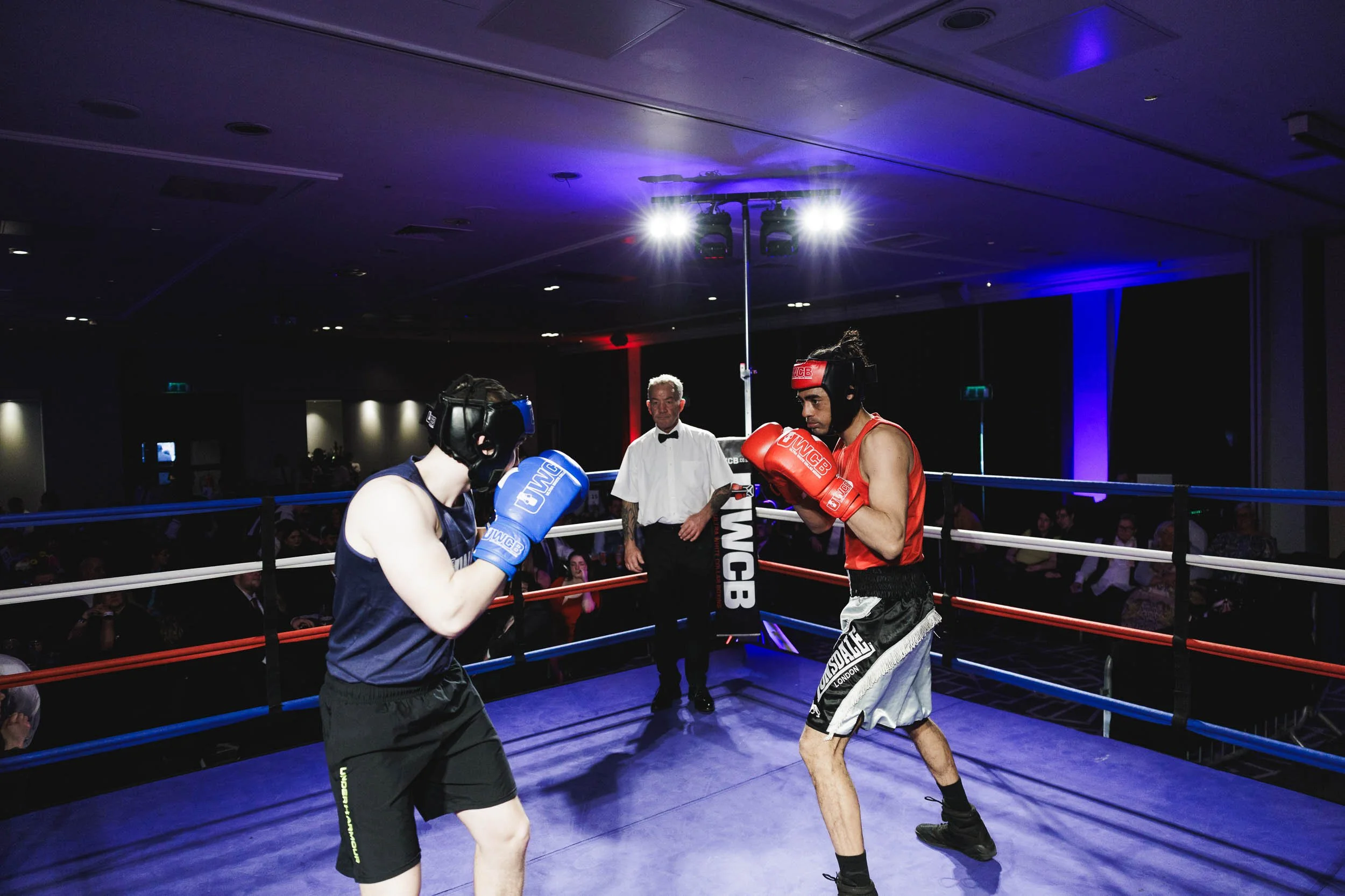 Two young men are sparring in a boxing ring, wearing protective headgear, gloves, and athletic clothing, while an official observes from inside the ring.