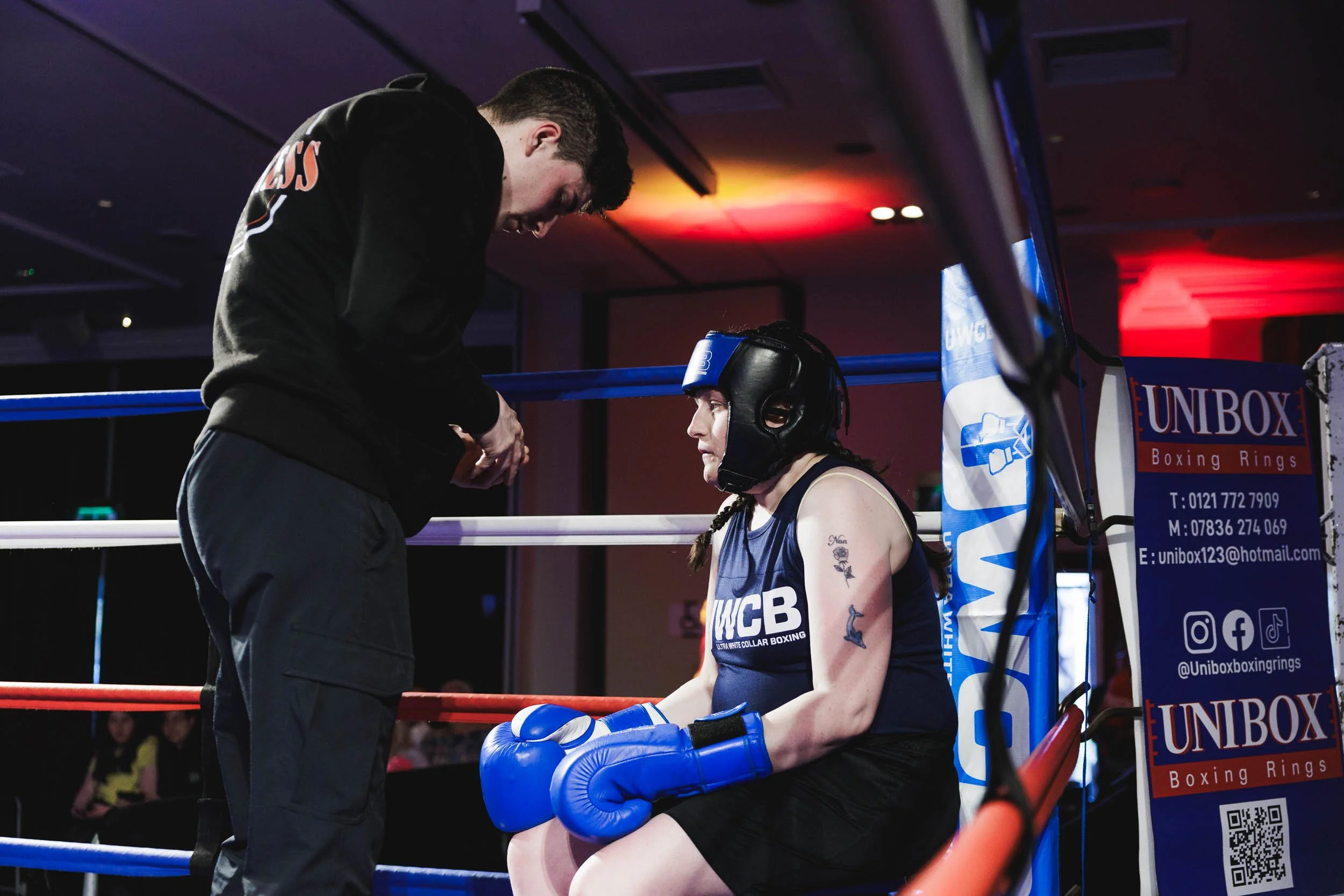 Female boxer sitting on the corner of the boxing ring with blue gloves and headgear, talking to her coach inside a boxing ring during a match or training session.