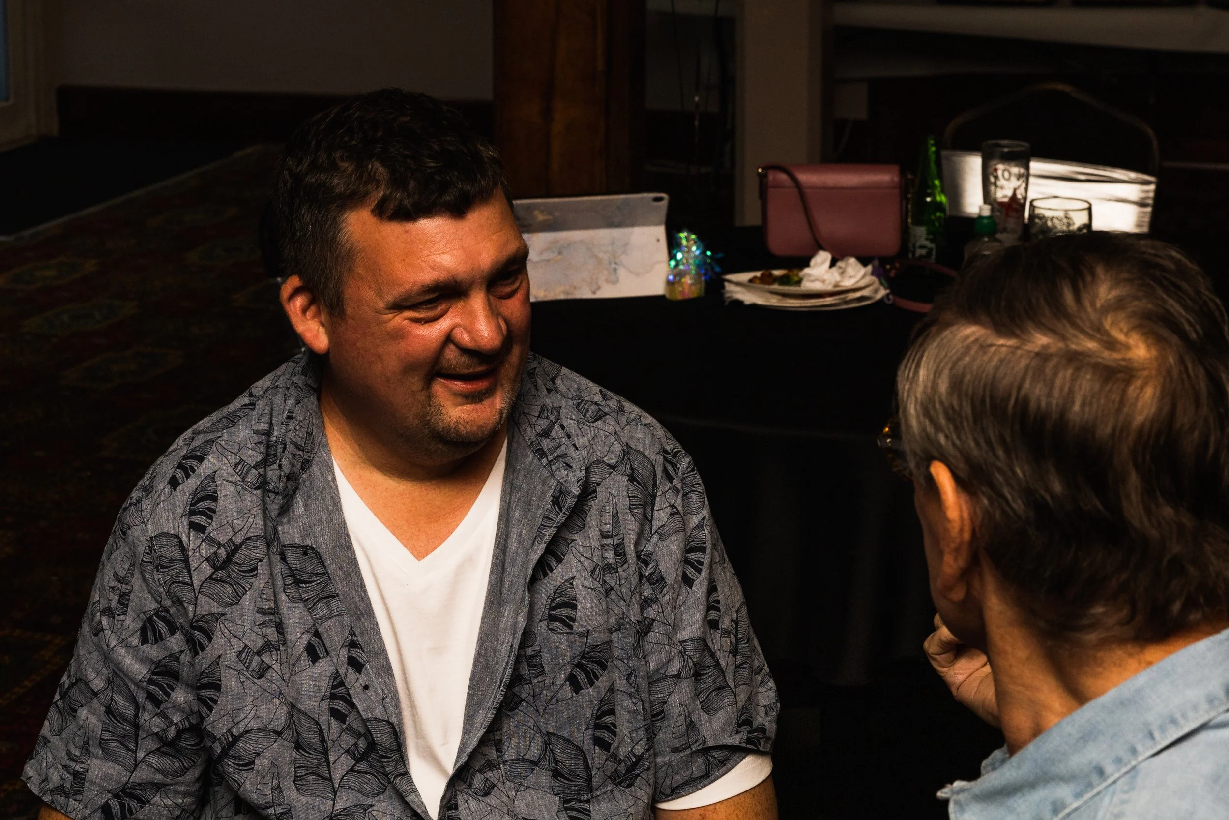 Two men engaging in conversation indoors, one smiling, with a table of plates and drinks behind them.
