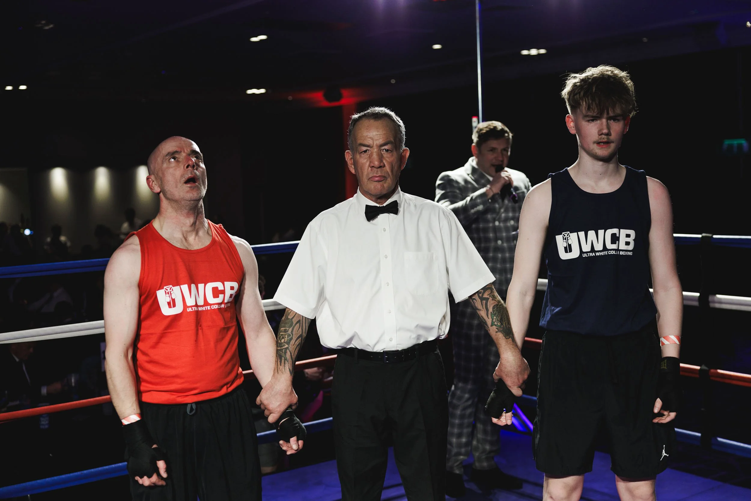 A boxing match ends with the referee holding the hands of the losing boxer in a red shirt and the winning boxer in a blue shirt, both standing in the ring, while a man with a microphone in the background announces the results.