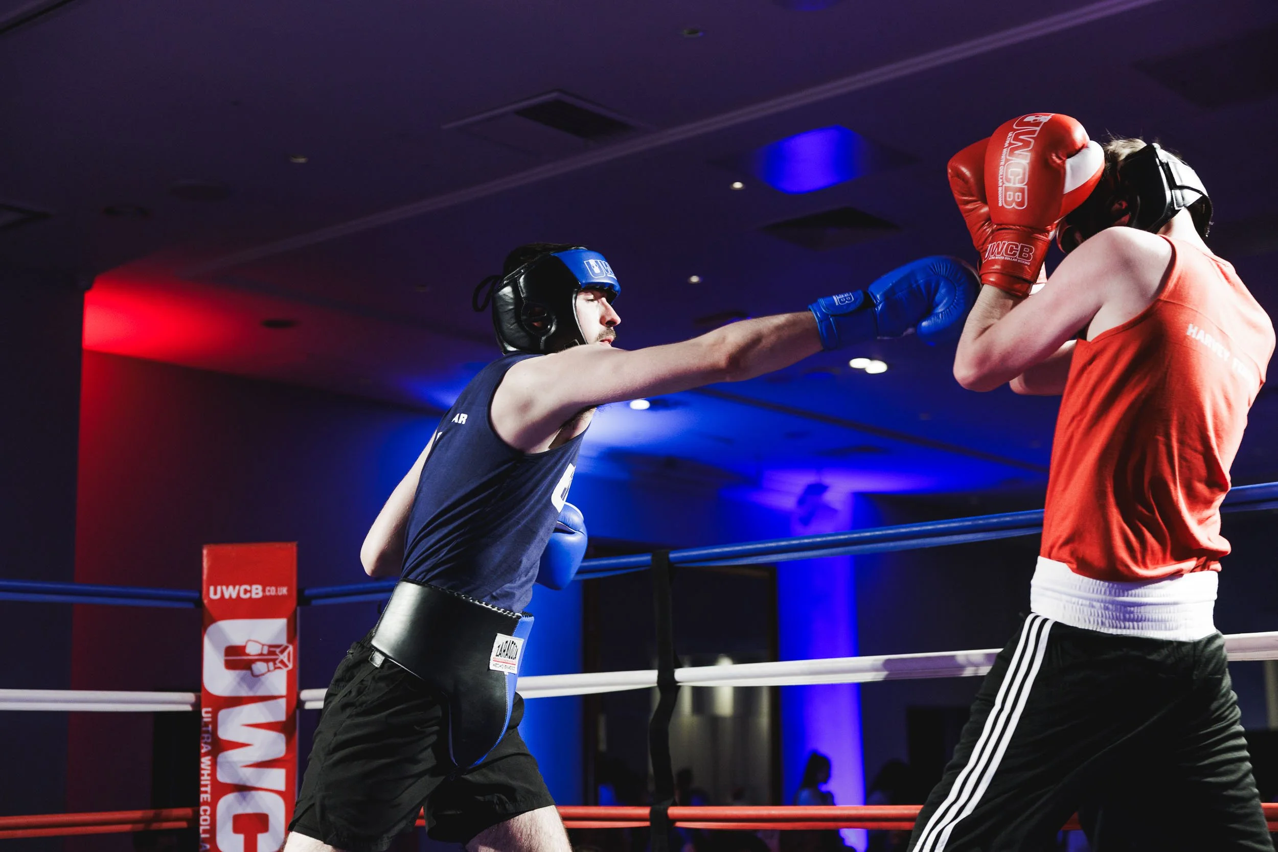 Two amateur boxers sparring in a boxing ring, one delivering a punch to the other's face, both wearing protective headgear and gloves, illuminated by colorful lighting.
