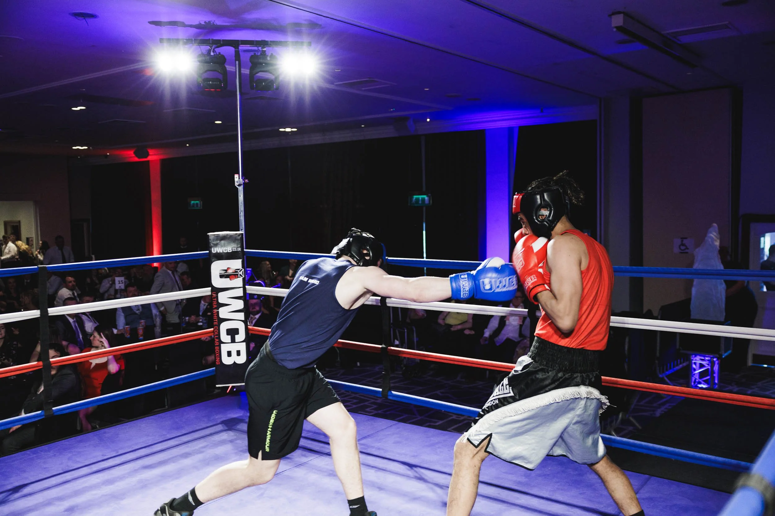 Two amateur boxers, one in red and the other in blue, sparring in a boxing ring with an audience watching in the background.