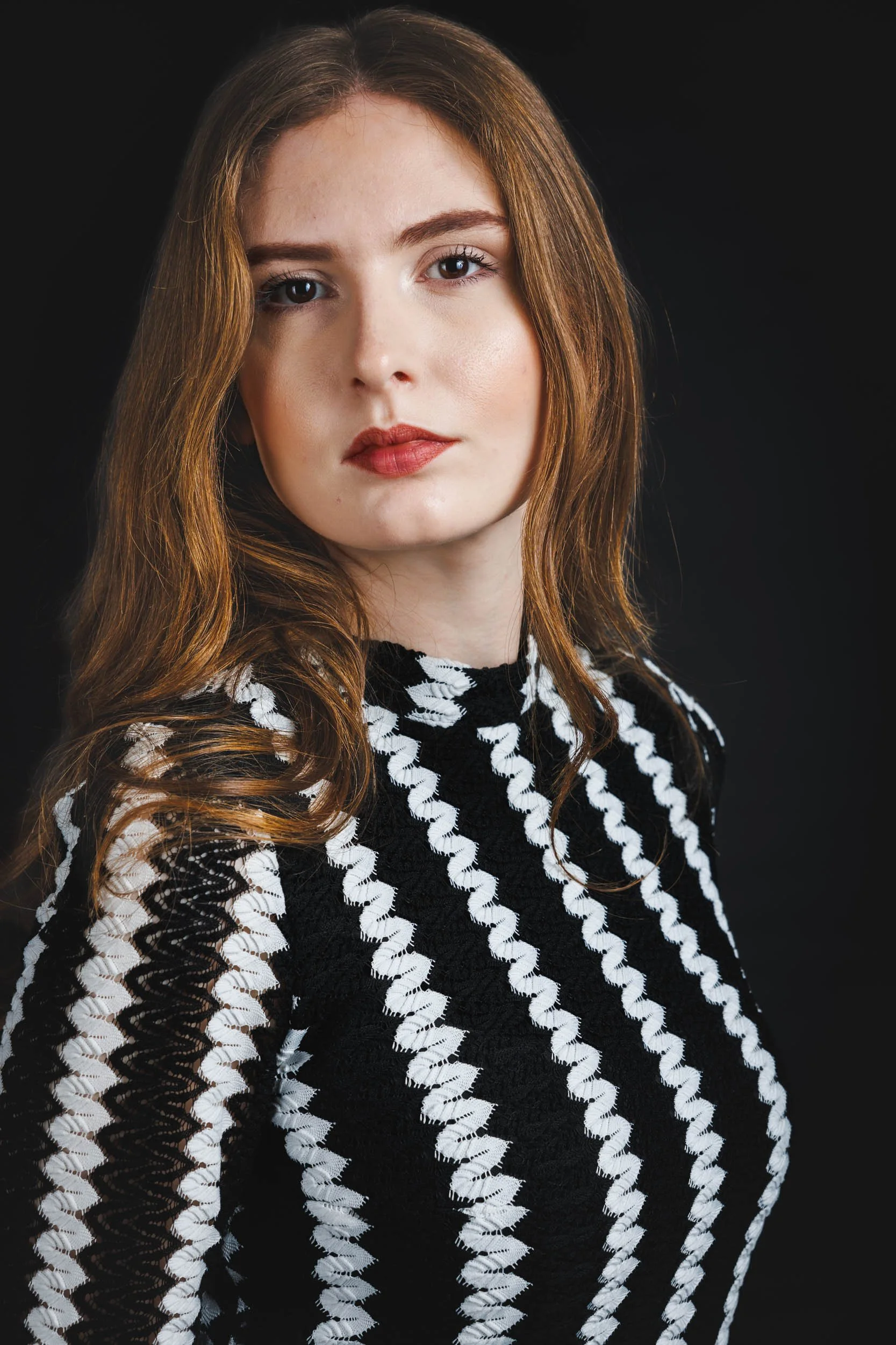 A young woman with long, wavy red hair and fair skin, wearing a black and white patterned top, looking directly at the camera against a black background.