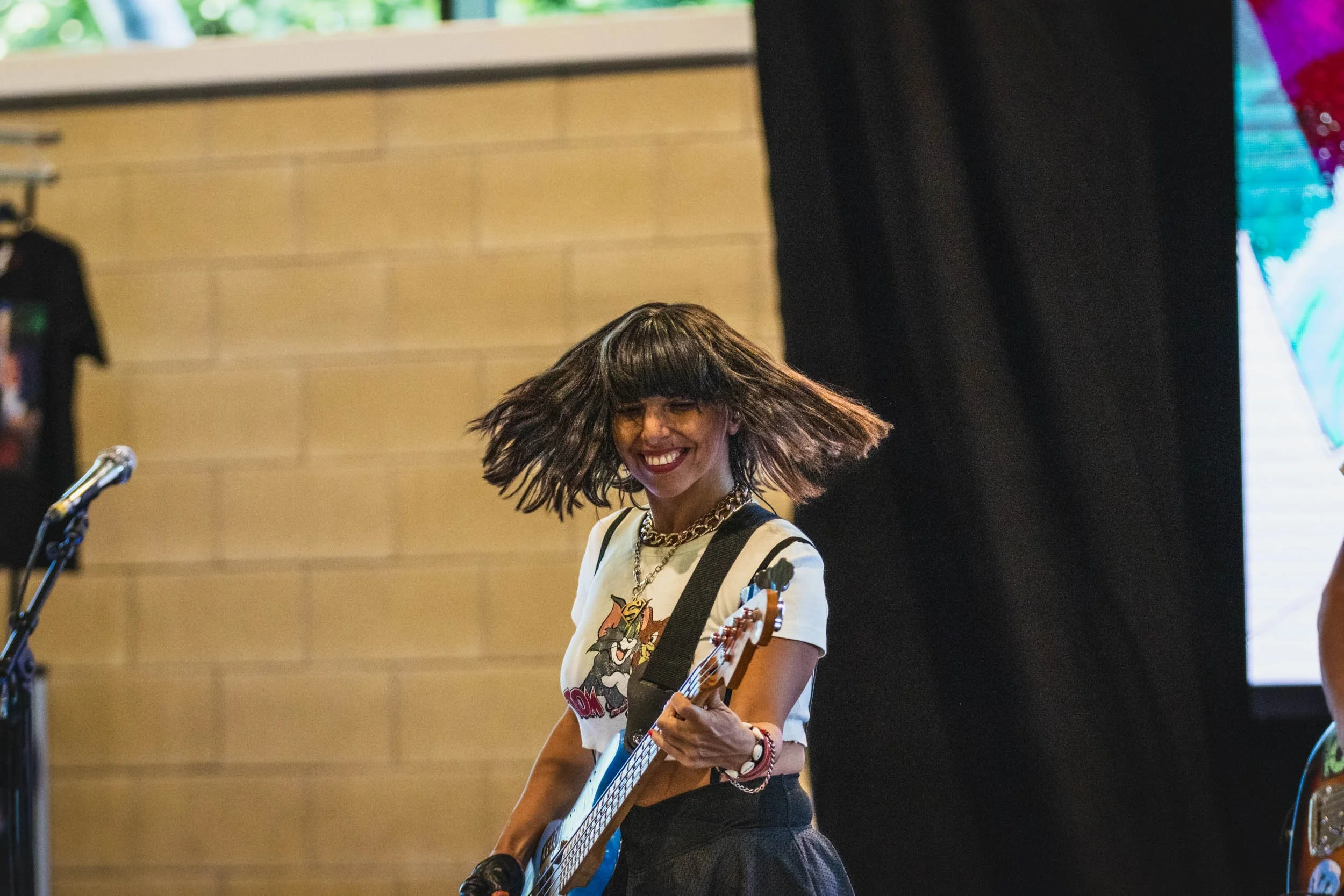 A woman with brown hair, wearing a white t-shirt with cartoon characters, playing an electric guitar and smiling at a live performance.