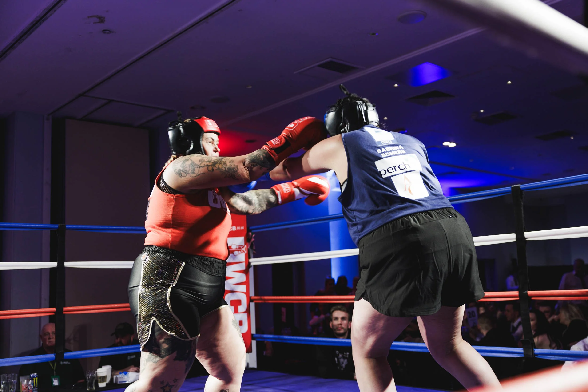 Two female boxers wearing protective headgear and gloves engaged in a boxing match inside a ring, with one delivering a punch to the other.