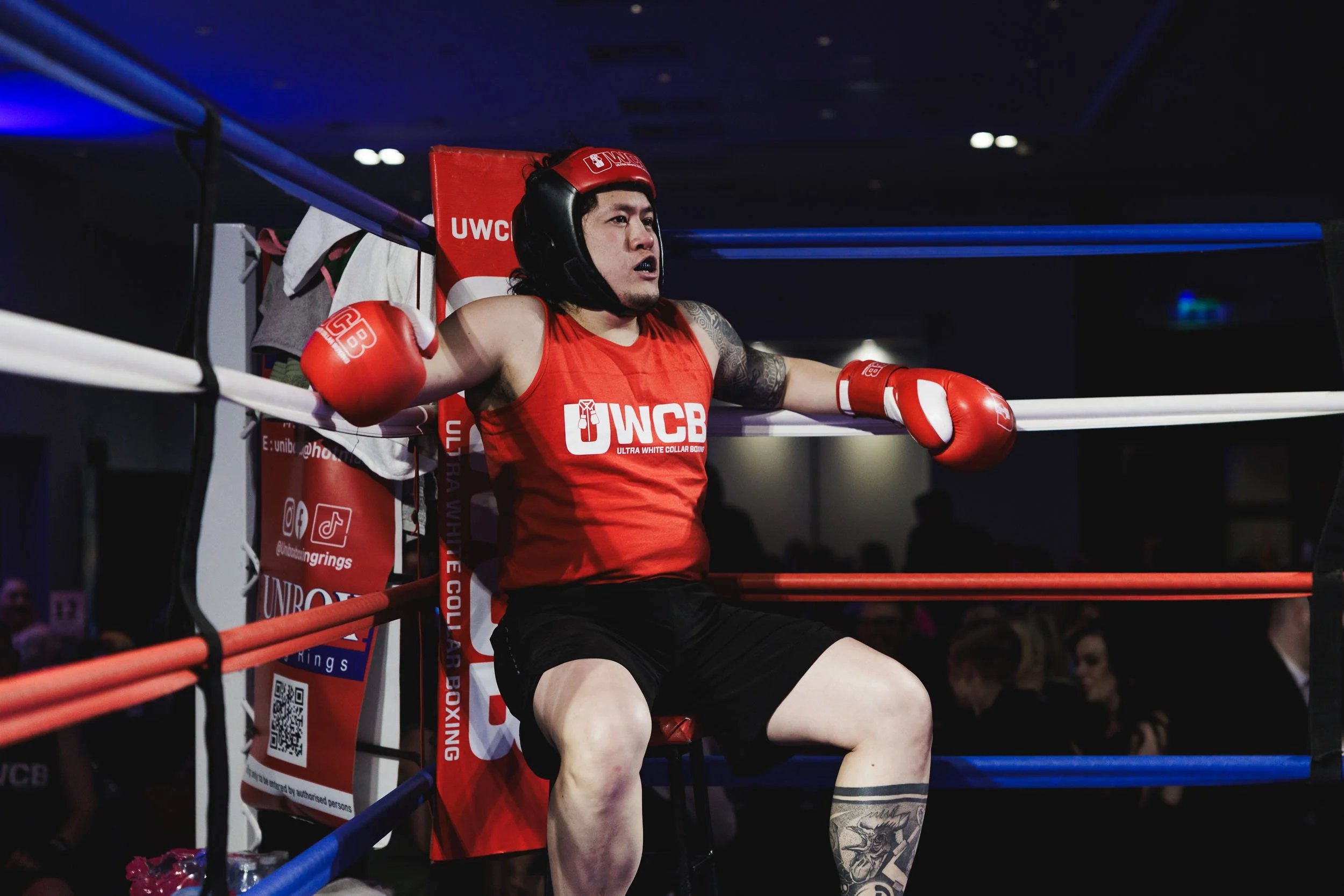 A male boxer sits on a chair outside the boxing ring, wearing red gloves, a red tank top with 'UWCB' logo, and a protective helmet, after a match.