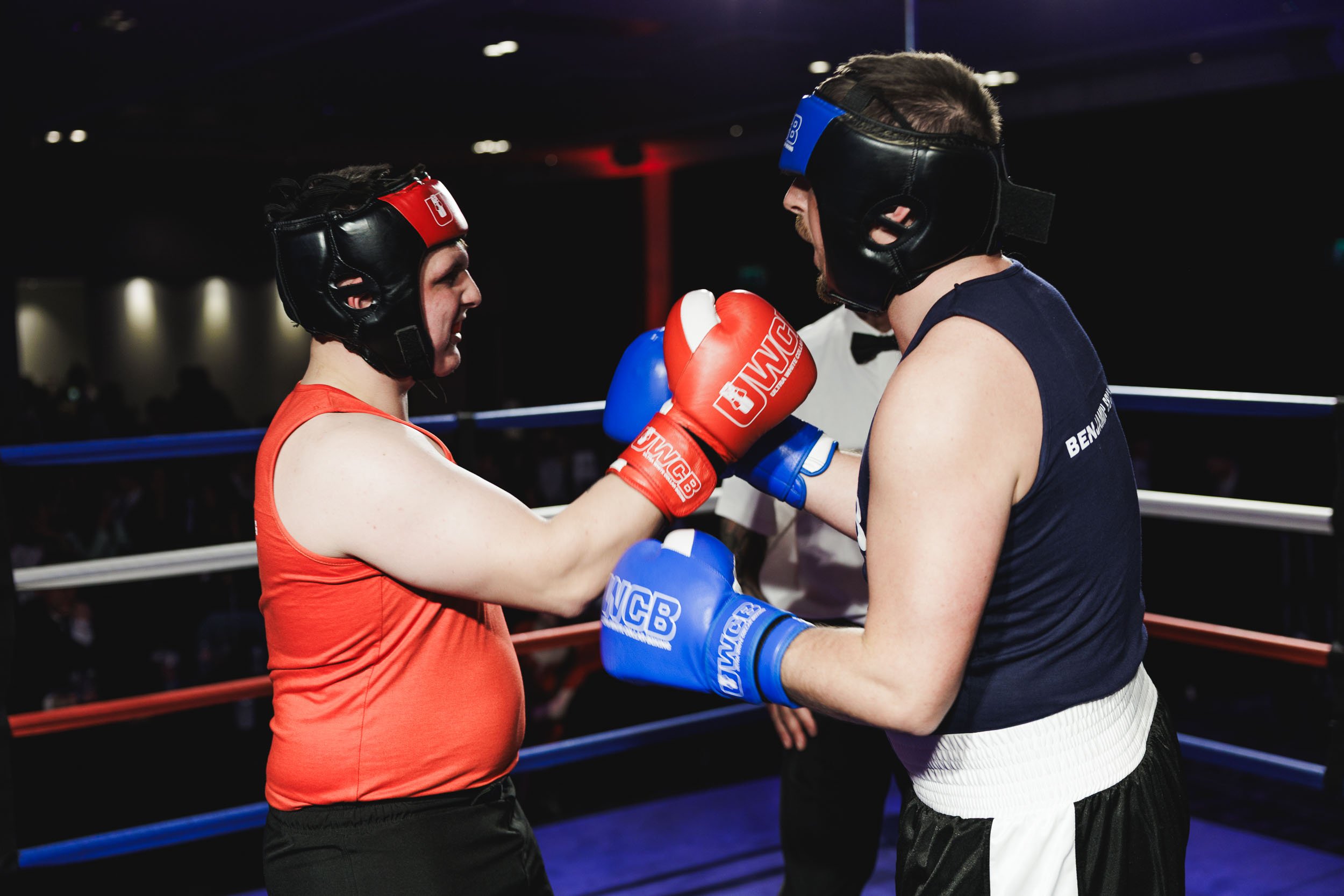 Two female boxers wearing protective headgear and gloves, one in red and the other in blue, face each other inside a boxing ring, engaged in a match.