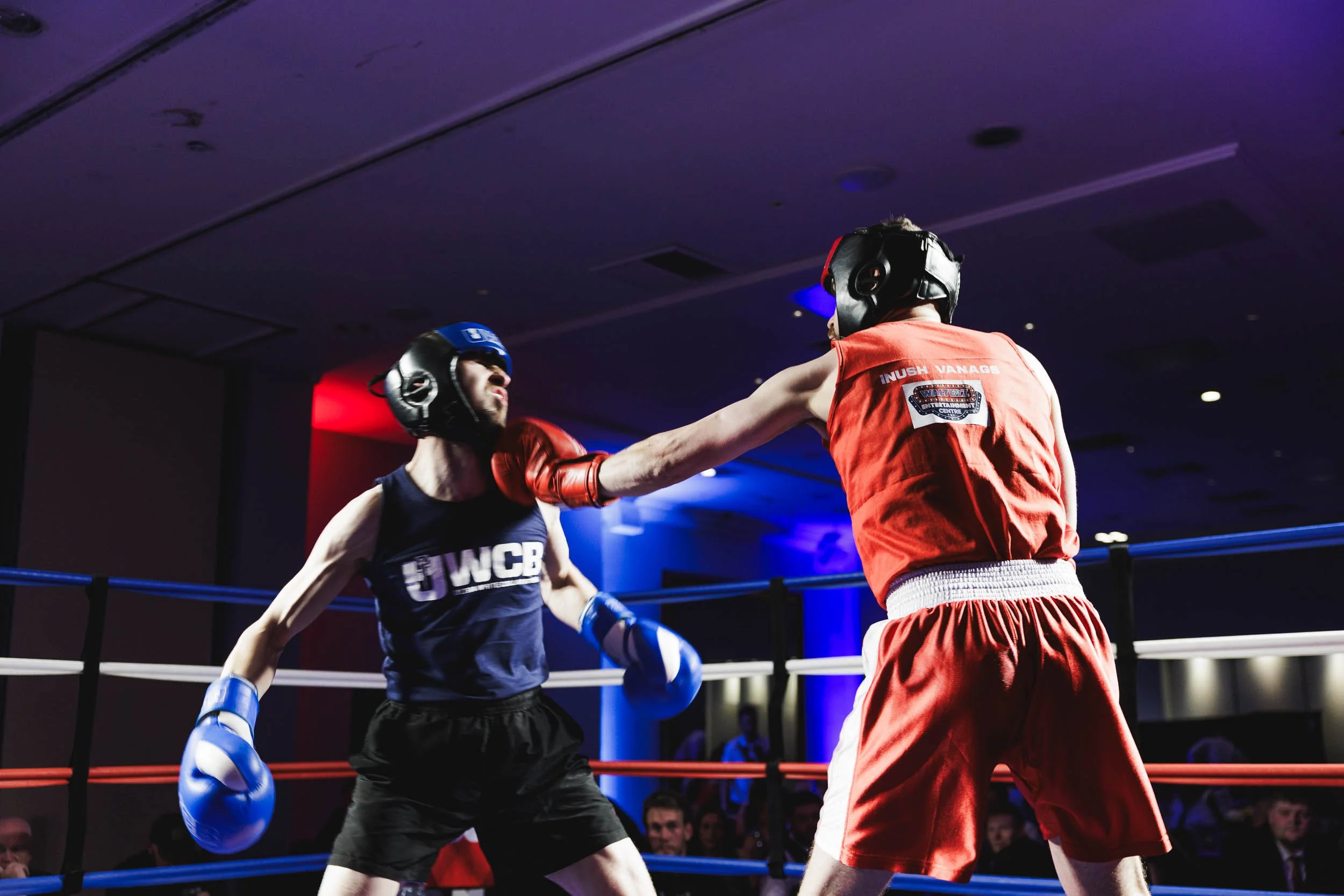 Two male boxers in a boxing ring engaged in a fight. One boxer in red is delivering a punch to the face of the other boxer wearing black. Both are wearing protective headgear and boxing gloves. The background shows audience members watching the match