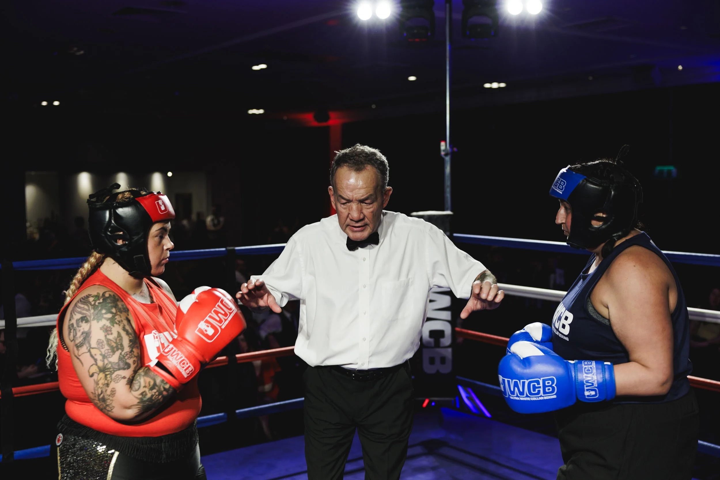 Two female boxers with protective headgear and gloves stand in a boxing ring facing each other as a referee speaks between them.