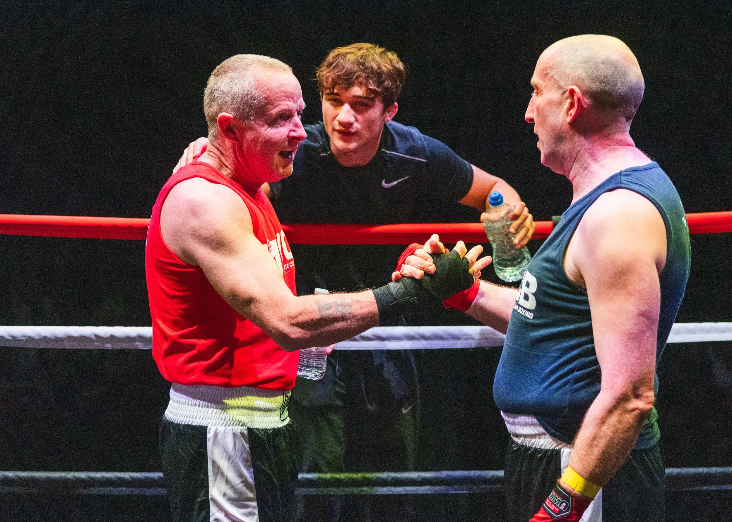 Two boxers shake hands inside a boxing ring, one wearing a red sleeveless shirt and the other in a dark sleeveless shirt, with a third person in the background holding a water bottle.