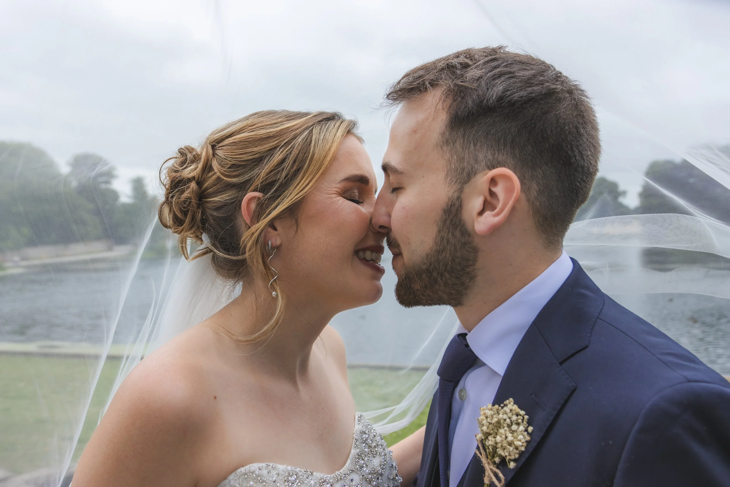 A bride and groom about to kiss outdoors near a body of water, with overcast skies and trees in the background.
