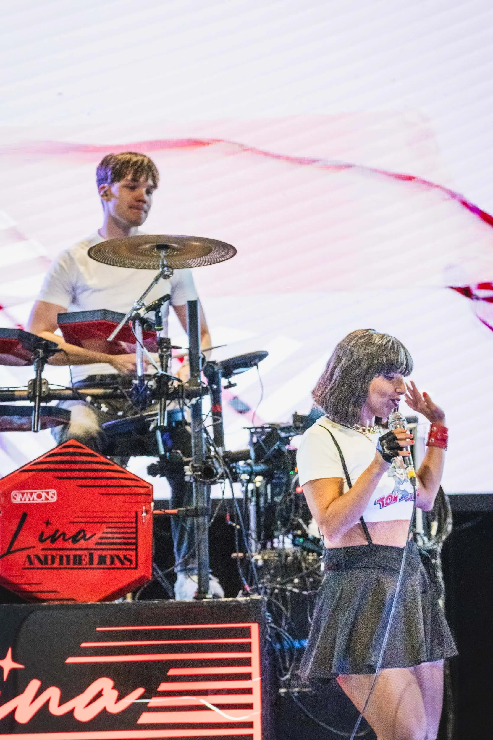 A young female singer with black hair and a white crop top singing into a microphone, performing on stage with a drummer playing electronic drums behind her.