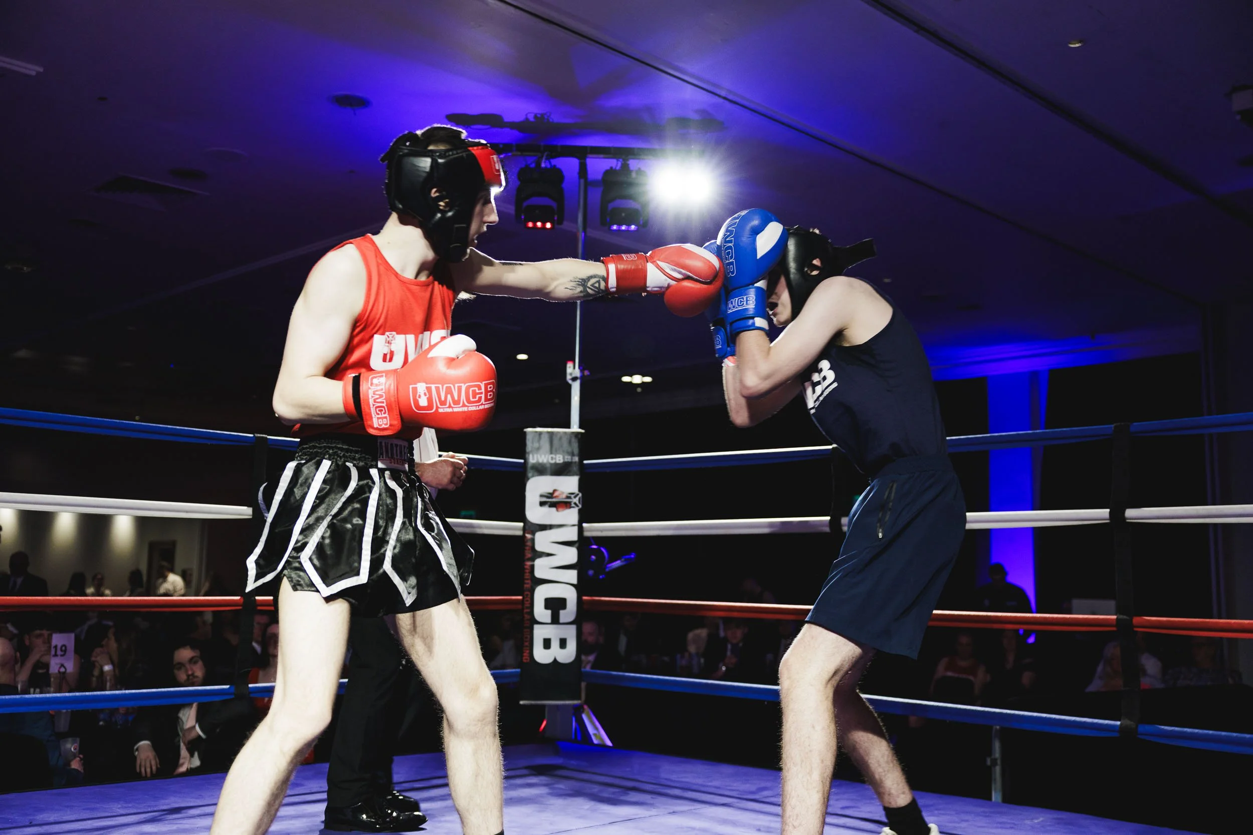 Two male young boxers with protective helmets sparring in a boxing ring, one delivering a punch, with spectators watching in the background.