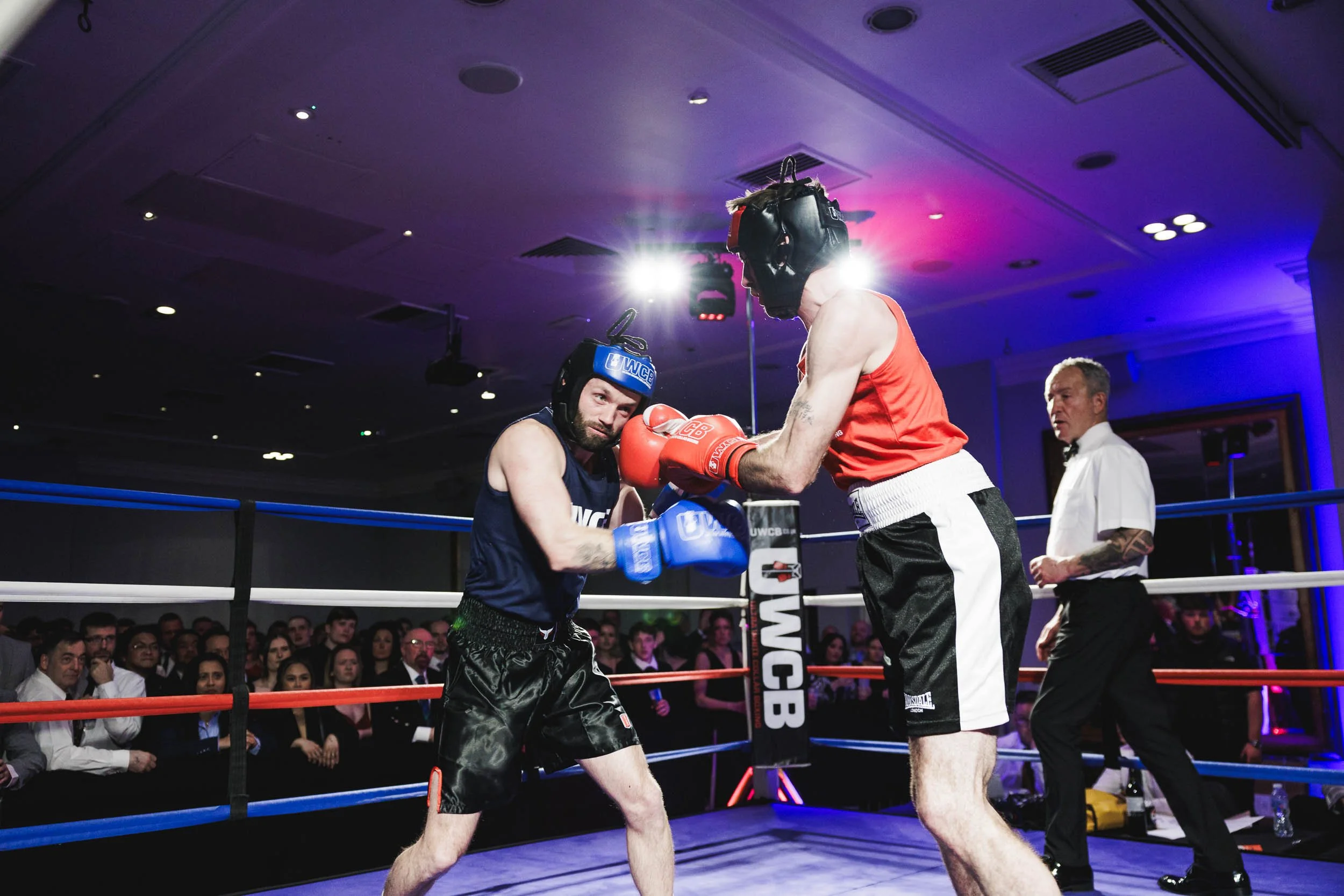 Two male boxers in a boxing ring are engaged in a match, with one in a red tank top and black shorts throwing a punch at the other, who is in a navy blue tank top and black shorts. They are wearing headgear and boxing gloves. An audience watches from
