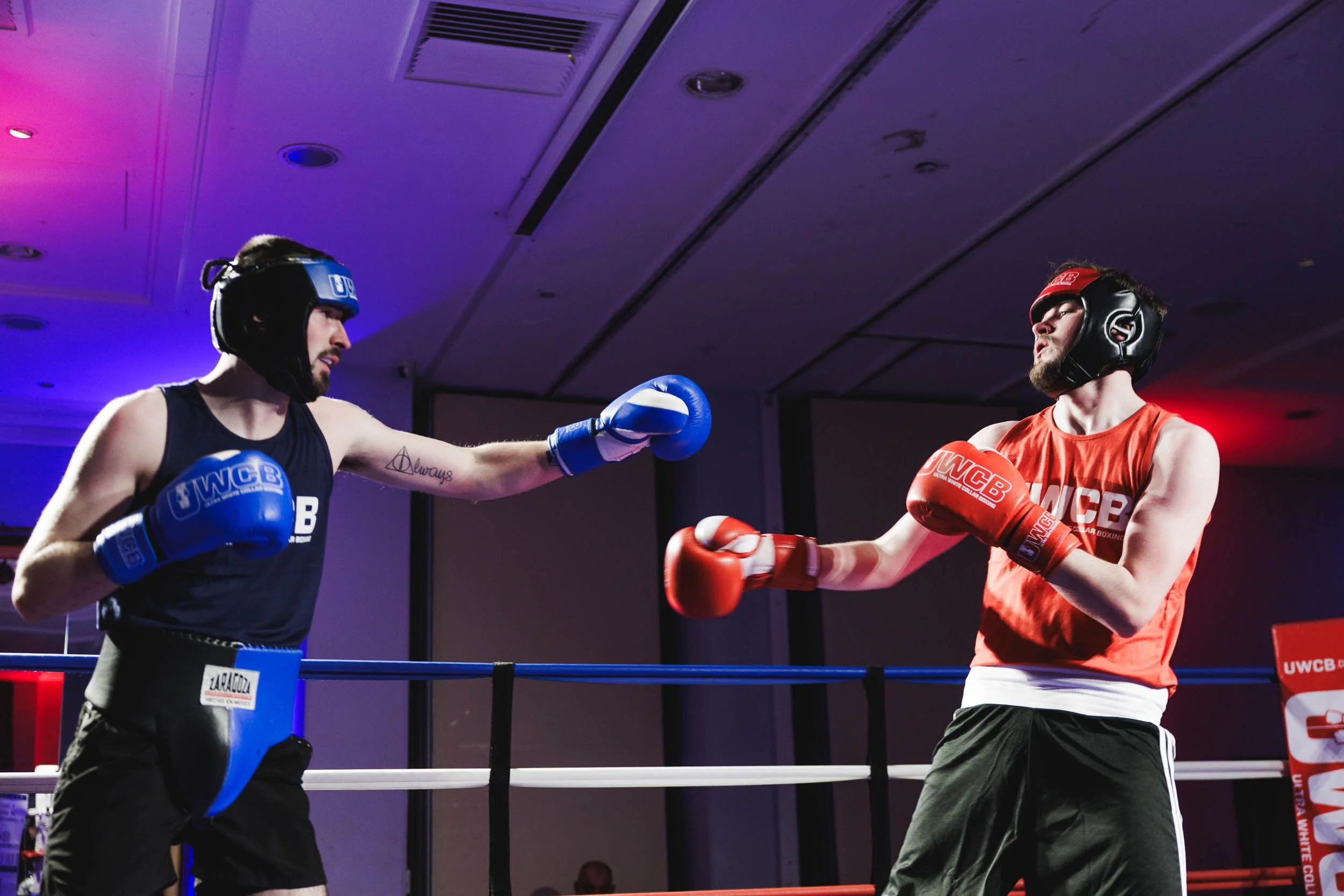 Two male boxers wearing headgear and gloves sparring in a boxing ring. One in black and the other in red, mid-punch exchange. Dim indoor lighting with blue and red lights.