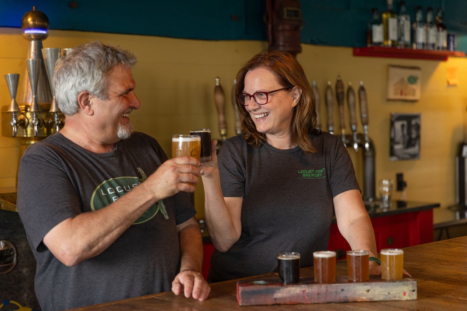 A man and a woman at a bar, smiling and clinking glasses of beer together. The man has gray hair and a beard, and the woman has red hair and is wearing glasses. There are five small glasses of beer on the bar in front of them.