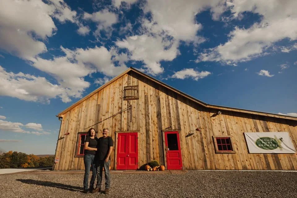 A wooden building with red doors and windows in a rural setting under a blue sky with clouds, featuring two women standing in front surrounded by pumpkins.