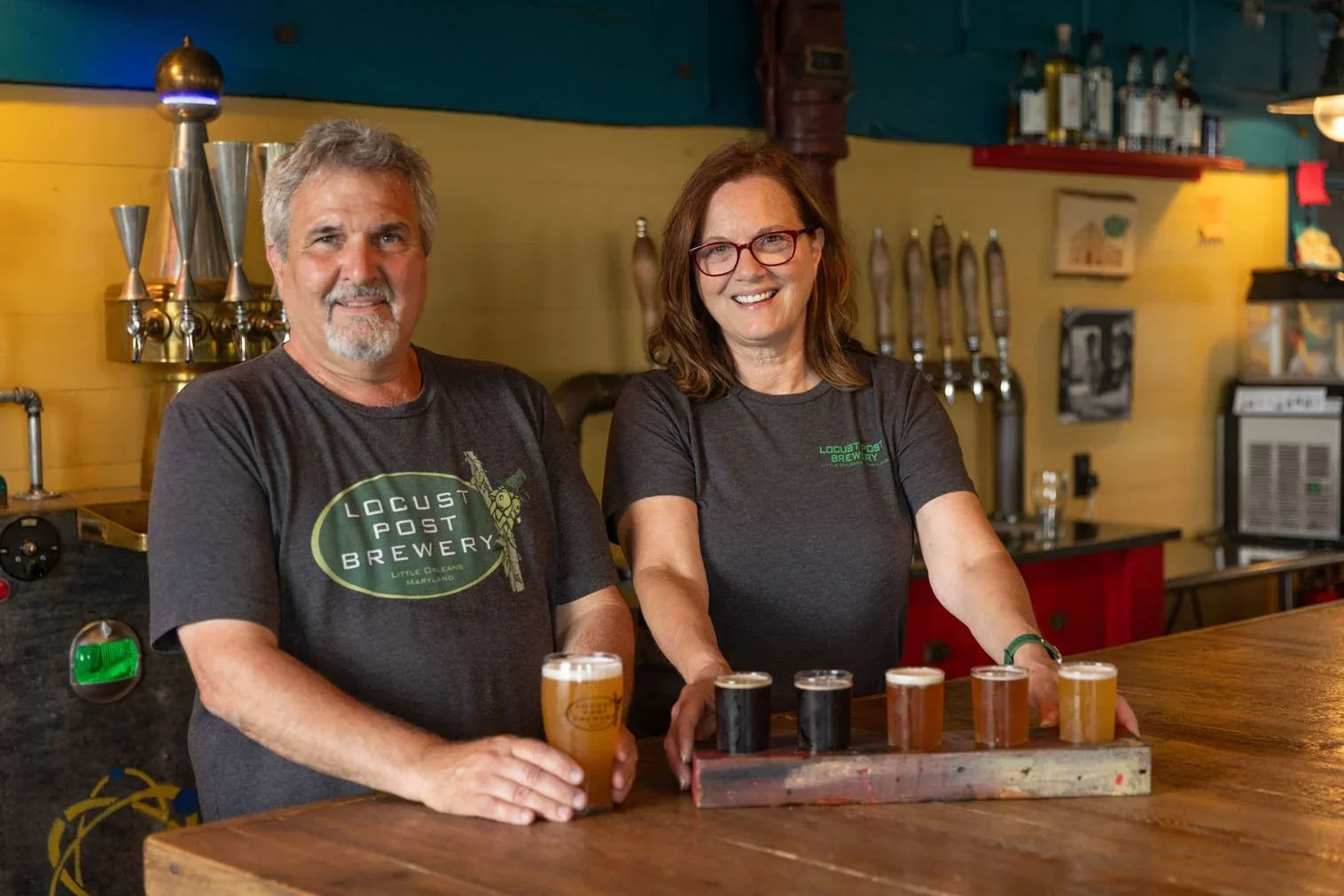 A man and woman standing behind a bar counter with six beer glasses, three filled with light beer and three with dark beer, in a brewery setting.