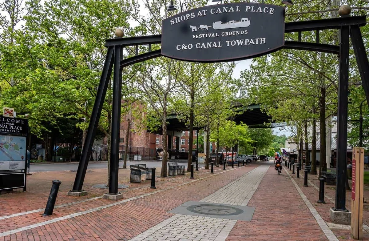 Entrance to Canal Place with a black sign that reads Shops at Canal Place, Festival Grounds, and C&O Canal Towpath, over a paved walkway lined with trees and benches.