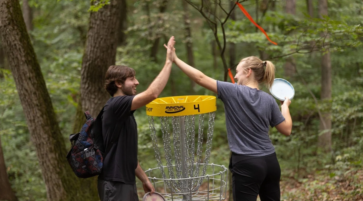 A man and woman high-fiving next to a disc golf basket in a forest, with the woman holding a disc in her hand.