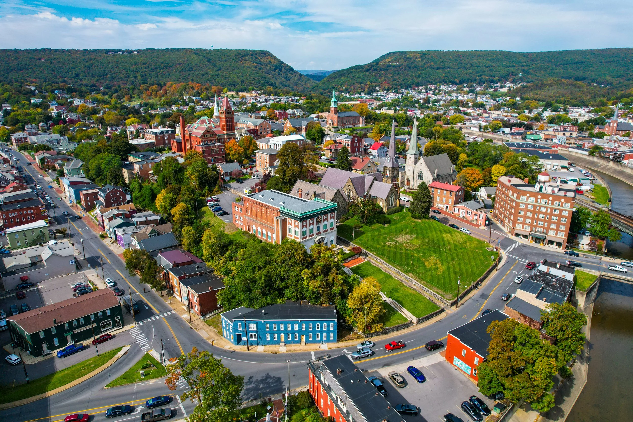 Aerial view of a small town with a mix of historic and modern buildings, surrounded by green hills and a river running through the area on a sunny day.