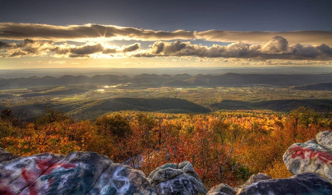 A scenic view of a valley with autumn-colored trees, distant mountains, and a dramatic sky with dark clouds, seen from a rocky overlook.