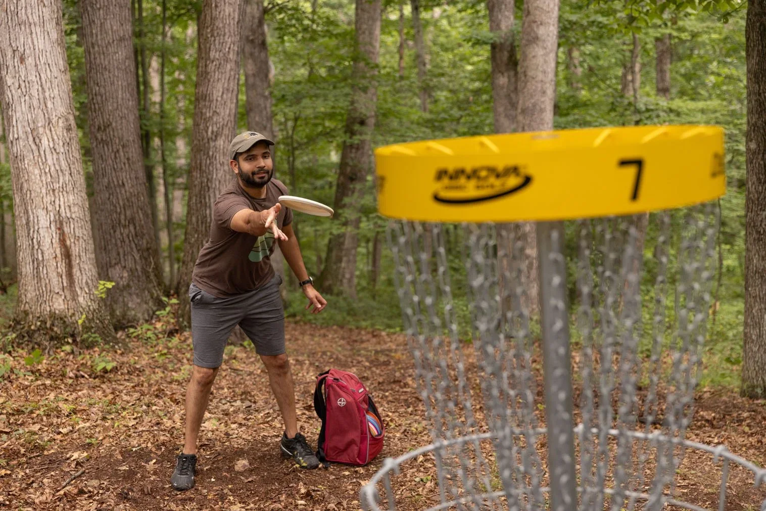 A man playing disc golf in a wooded area, reaching out to throw a disc towards a yellow disc golf basket with chains, with a red backpack on the ground beside him.