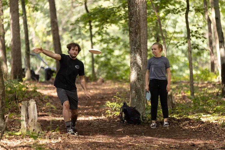 A man and woman playing disc golf in a wooded park. The man is throwing a frisbee and the woman is holding a disc. Trees and a backpack are visible in the background.