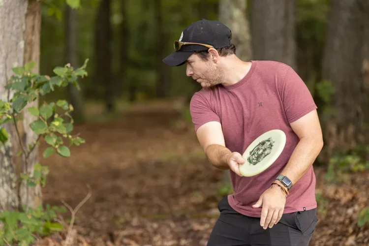 A man in a red t-shirt and black cap playing disc golf in a wooded area.