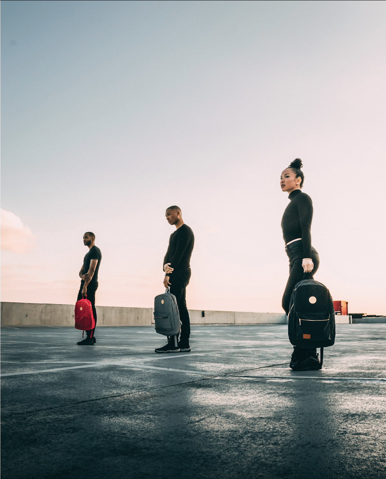 Three diverse individuals standing apart with backpacks on a rooftop parking lot during sunset, maintaining social distance.