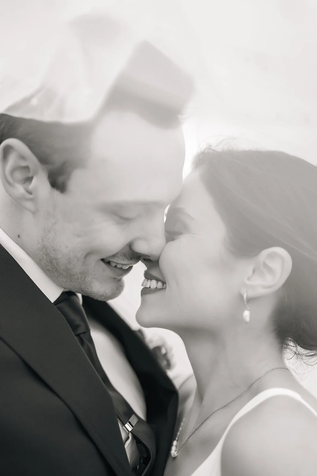 Black and white photo of a happy couple with foreheads and noses touching, smiling with eyes closed, dressed in formal wedding attire.